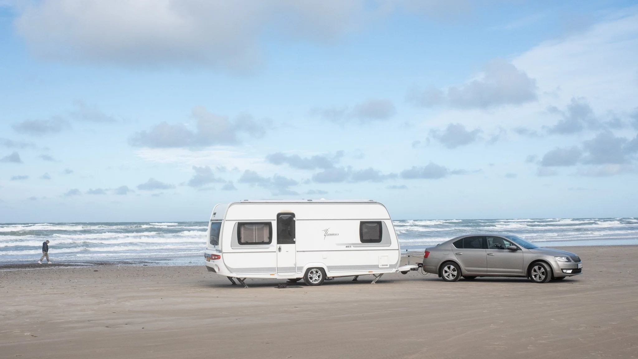 A beach scene with a white camper trailer, a silver car, and a person walking near the shoreline. The sky is partly cloudy, and waves are crashing on the shore.