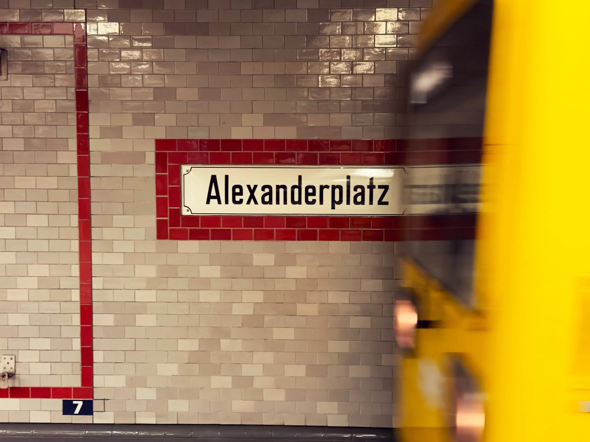 Underground subway station platform with tiled walls, a sign that reads "Alexanderplatz," and a partially visible yellow safety barrier in the foreground.