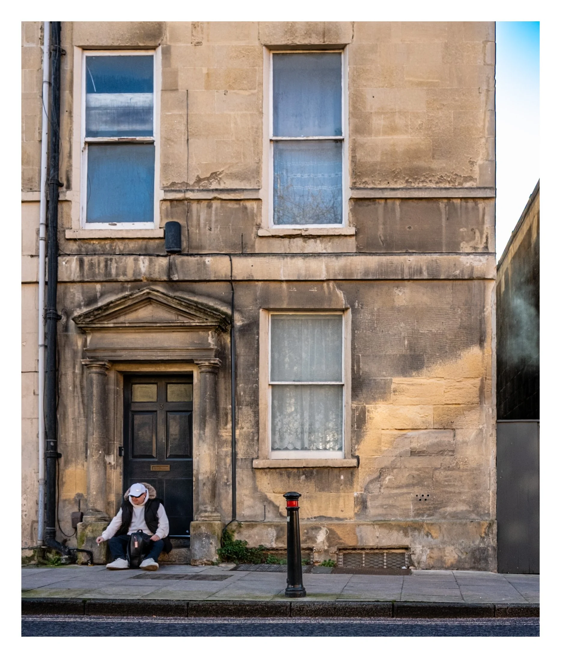 A man sitting on stone steps in front of an old stone building with four windows and a black door, wearing a white cap, white jacket, dark vest, and backpack.