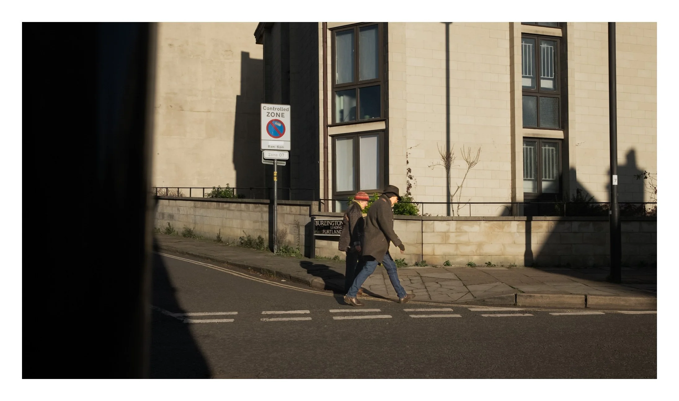 Two people walking across a crosswalk on a city street in front of a modern building with large windows. There are street signs, including a no parking zone sign, and a lamppost, with long shadows cast by the sunlight.