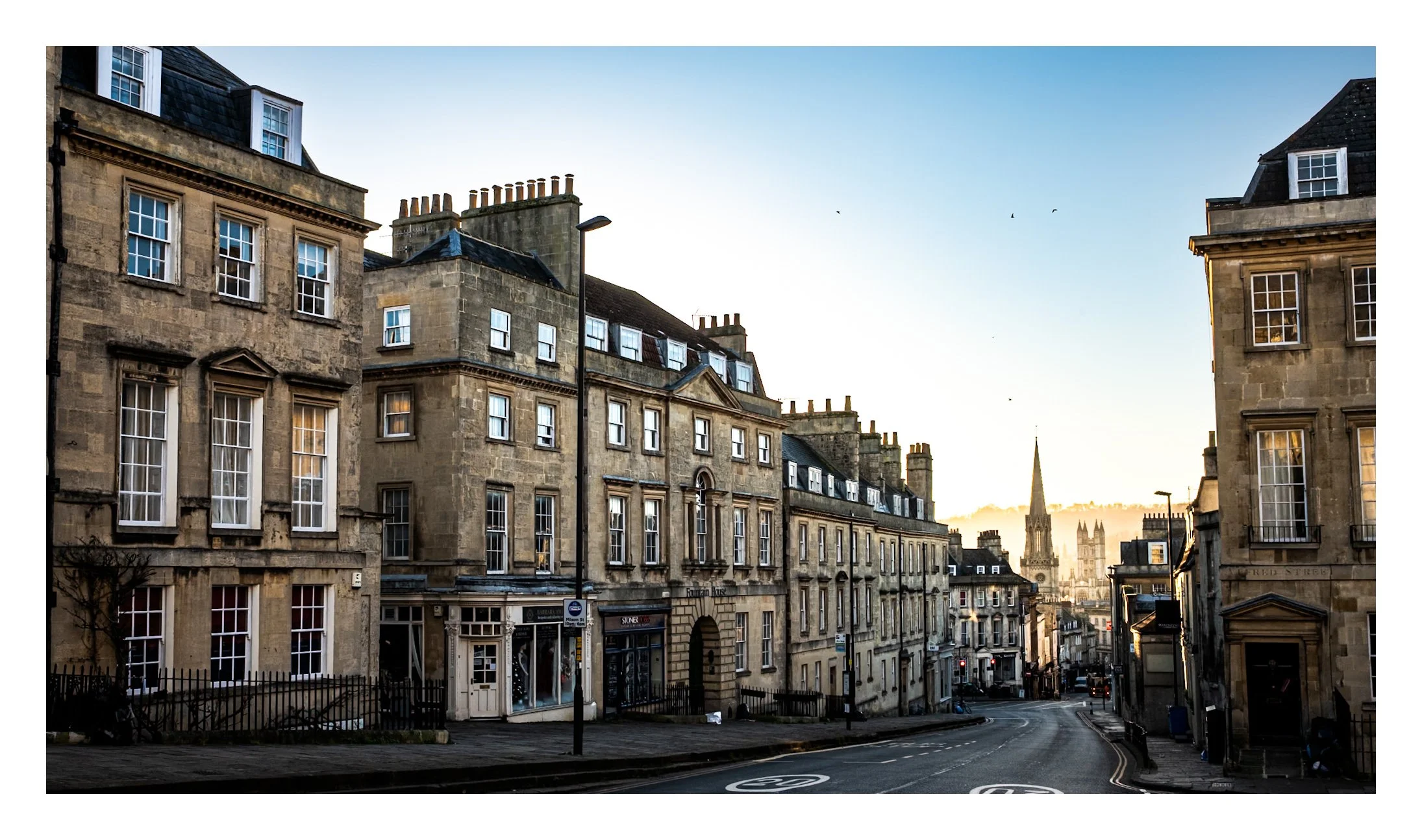 View of a street in a historic city with classic stone buildings and church spires in the background, captured during early morning or late afternoon.