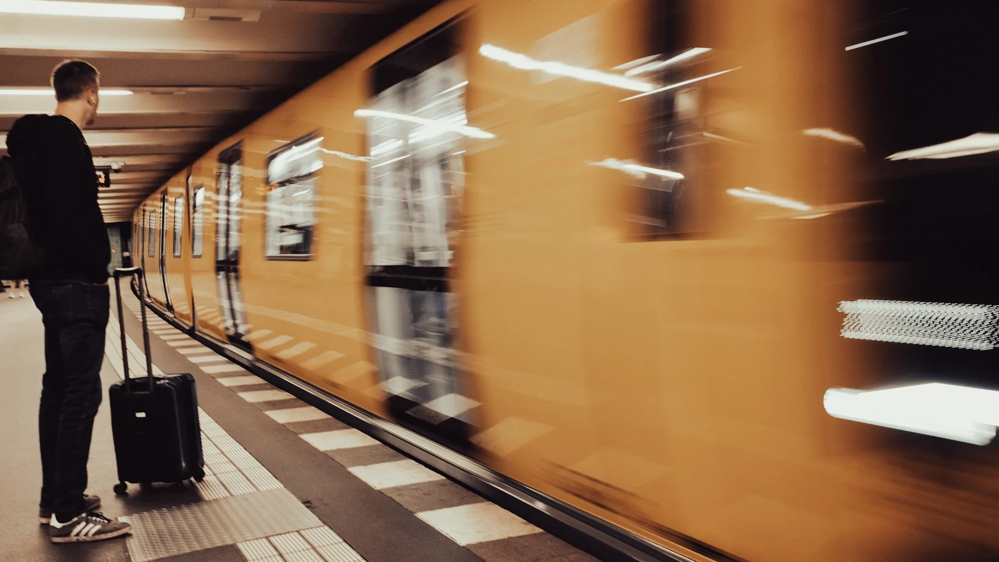 A person with a black suitcase standing on a subway platform next to a moving yellow train with blurred lights.
