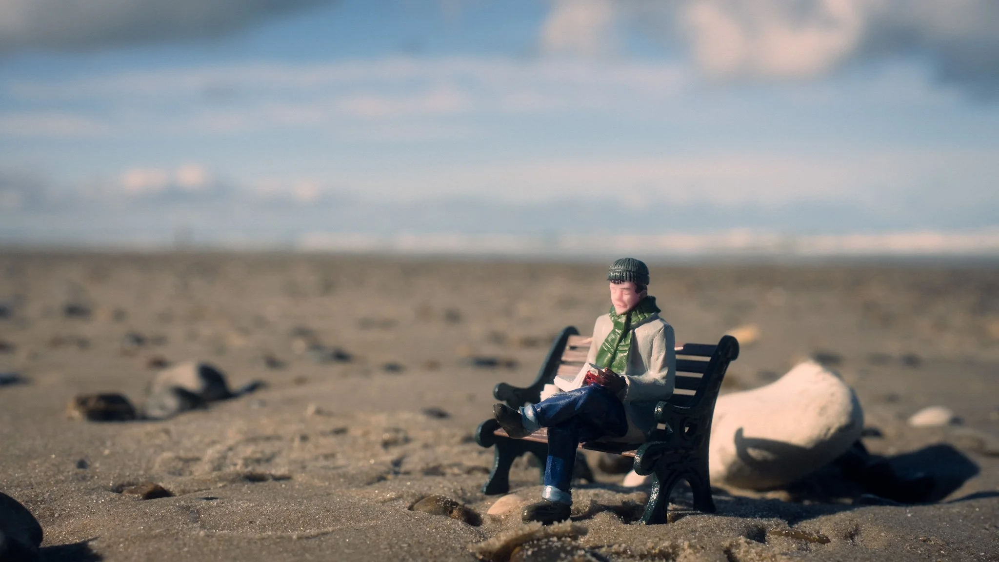 A miniature figurine of a man sitting on a park bench on a sandy beach, wearing a gray jacket, green scarf, and gray beanie, with rocks scattered on the sand and cloudy sky in the background.