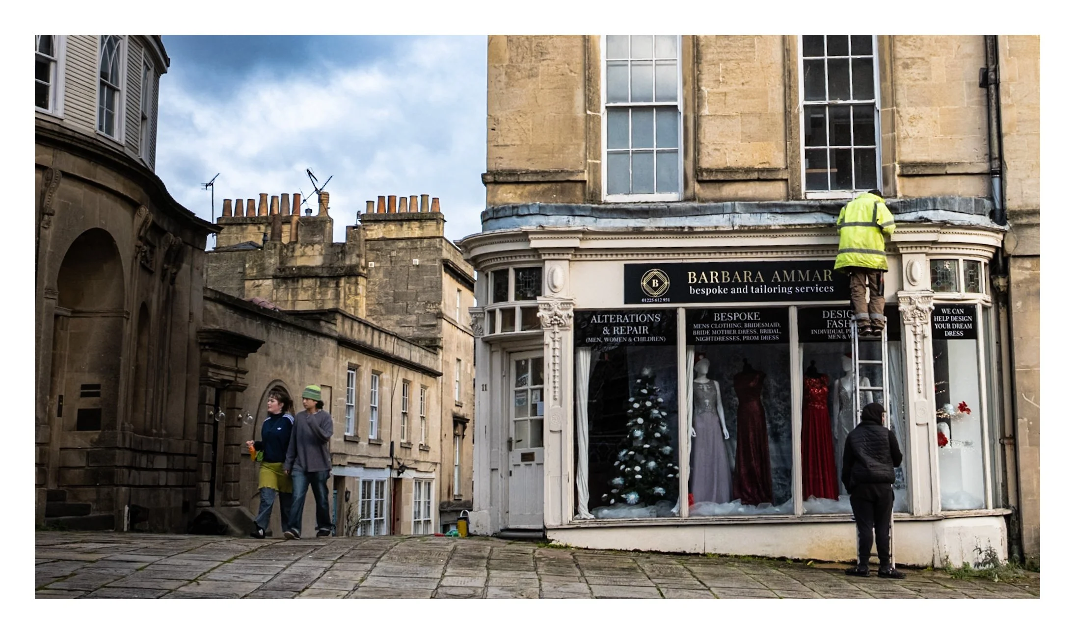 A street scene featuring a clothing store named Barbara Ammar with mannequins dressed in fancy clothes and a decorated Christmas tree in the window. A worker on a ladder is doing maintenance above the store. Two people are walking on the sidewalk, an