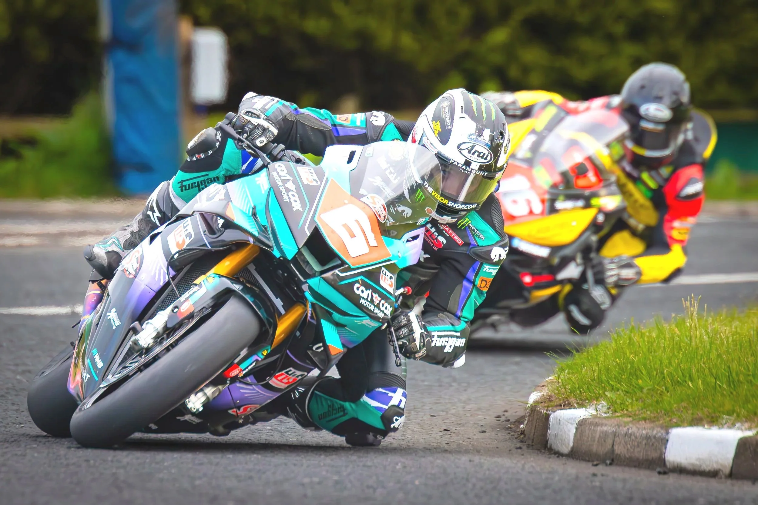Two motorcycle racers leaning into a turn on a racing track, with the rider in front wearing a teal, black, and purple suit and the rider behind in a red, yellow, and black suit.