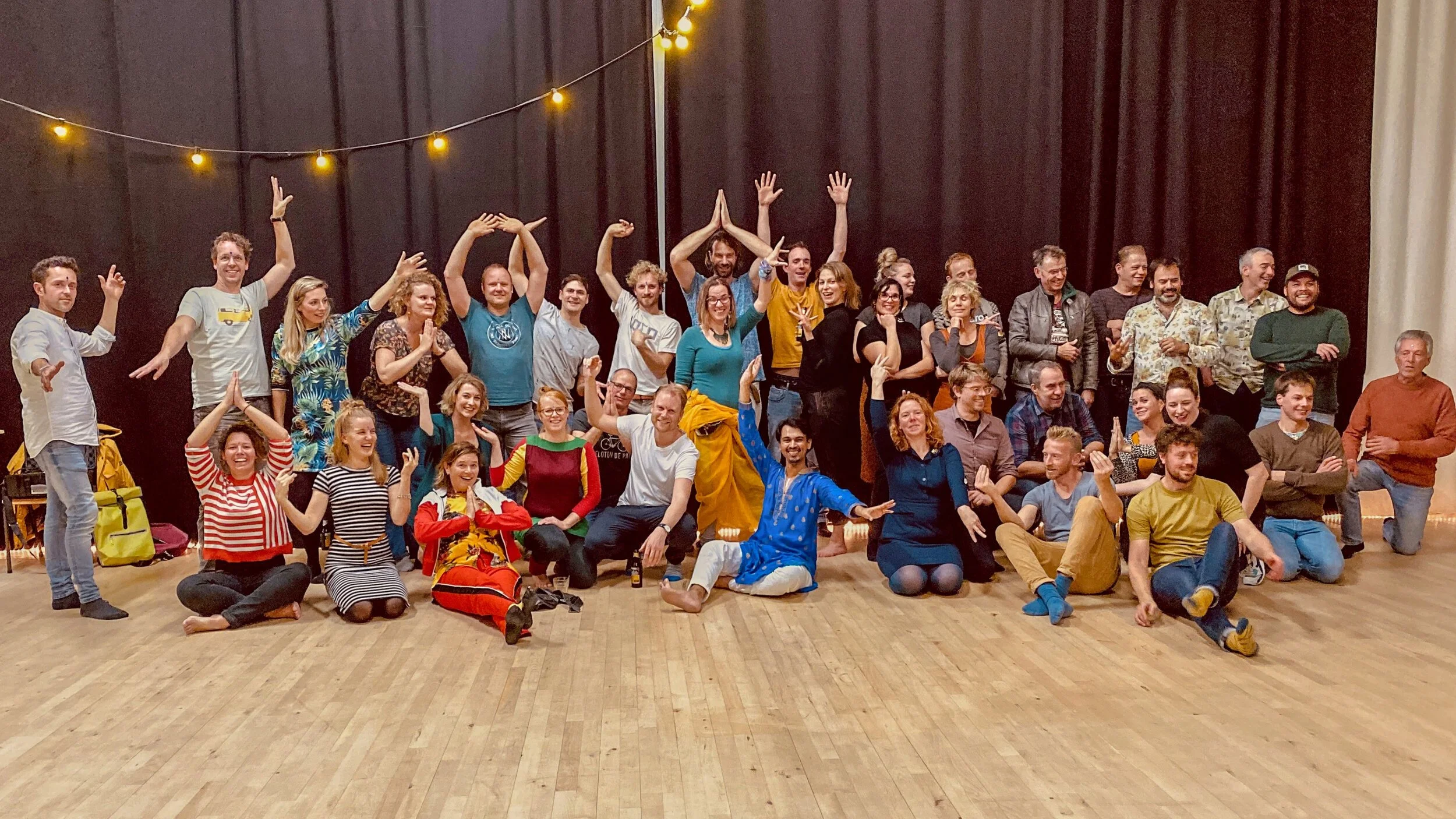 A large group of people posing indoors on a wooden floor, with playful gestures and smiles. They are in front of a black curtain with string lights, suggesting a casual, festive gathering or event.