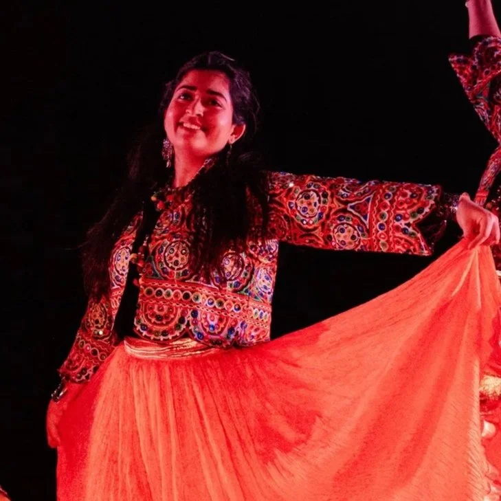 People dancing at an outdoor event, wearing colorful traditional costumes. A woman in the foreground is smiling and raising her arm. The stage in the background has lights and a canopy.