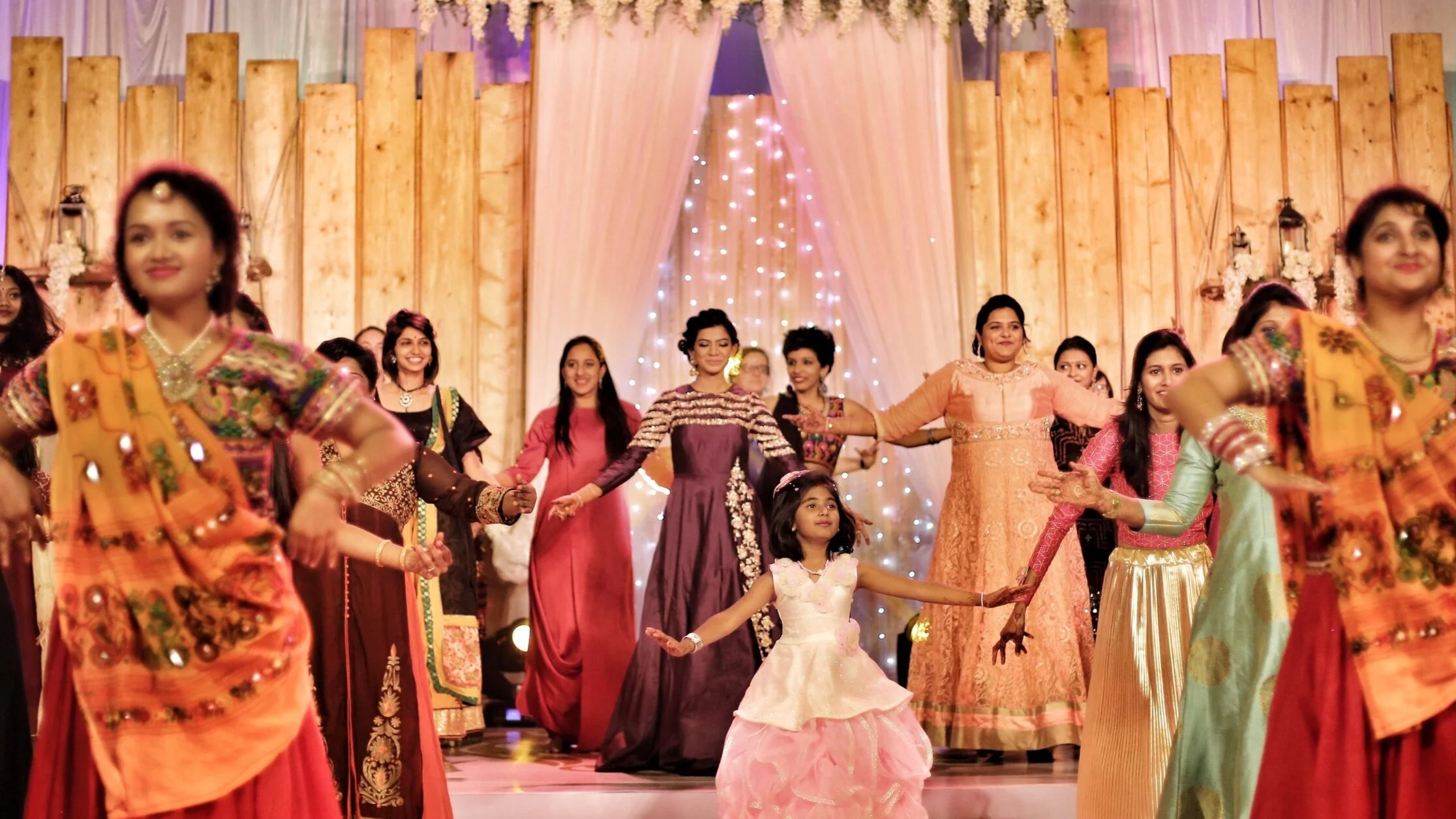 Group of people wearing colorful traditional Indian attire, dancing at an indoor event with decorative lights and wooden backdrop.