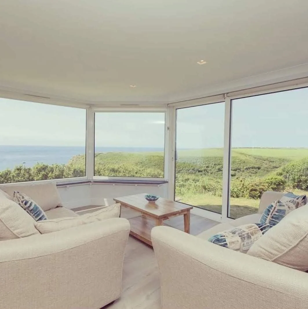 Sunroom with large glass windows showing a green landscape and ocean view, furnished with cream-colored sofas, patterned pillows, and a wooden coffee table.