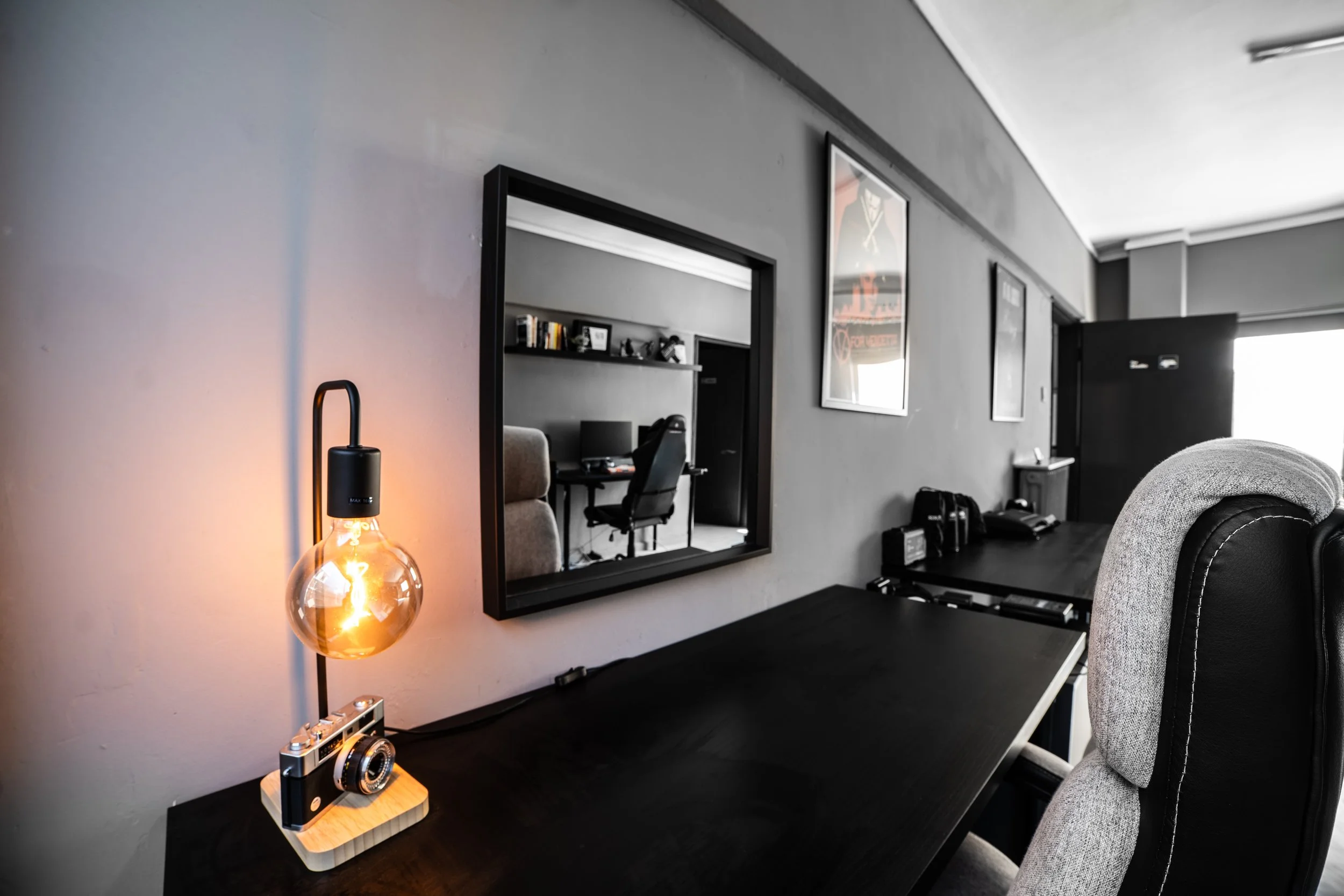 A black desk with a vintage camera on a wooden stand and a lit Edison-style bulb lamp nearby, against a gray wall with framed artwork and a large mirror reflecting an office space with a desk, chairs, and shelves.