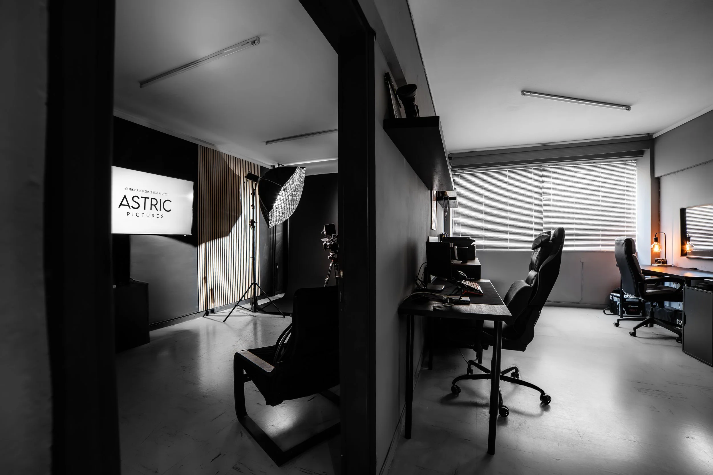 A darkened office with a large window covered by blinds, several desks with computers, black office chairs, and warm lighting from small lamps, with a production setup including a softbox and a monitor displaying "ASTRIC PICTURES".
