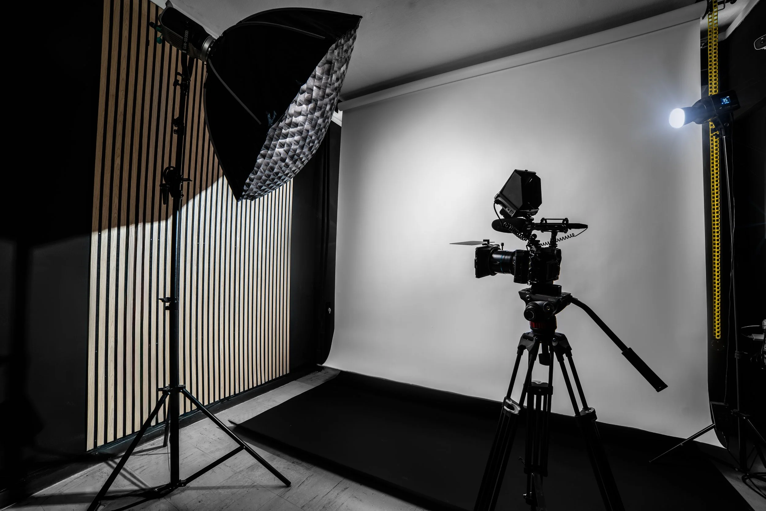 Photography studio setup with a white screen, professional camera on a tripod, and lighting equipment, including a large softbox, in a room with wooden wall paneling.