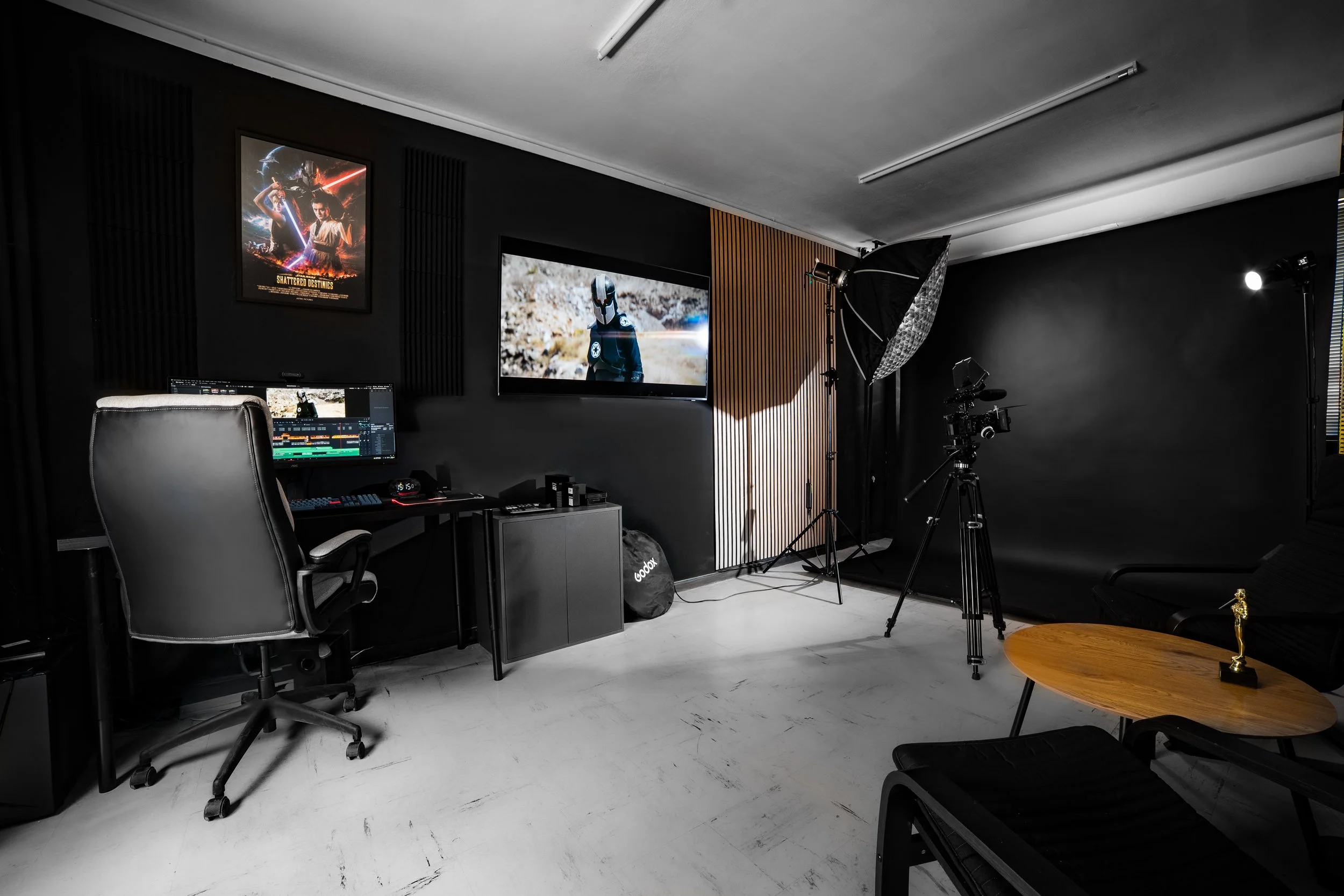 A professional photography studio setup with a black backdrop, a large monitor displaying a sci-fi character, studio lights, a camera on a tripod, computer workstation, and various filming equipment.