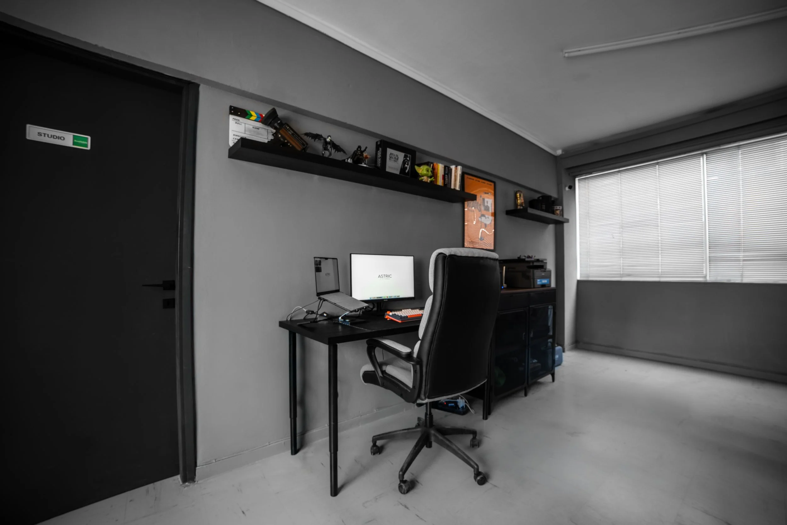 A workspace with a black desk and a black office chair, a computer monitor, a keyboard, and a mouse. Above the desk are two black floating shelves with books and figurines, including a dragon figure. The room has a window with closed blinds, and the 