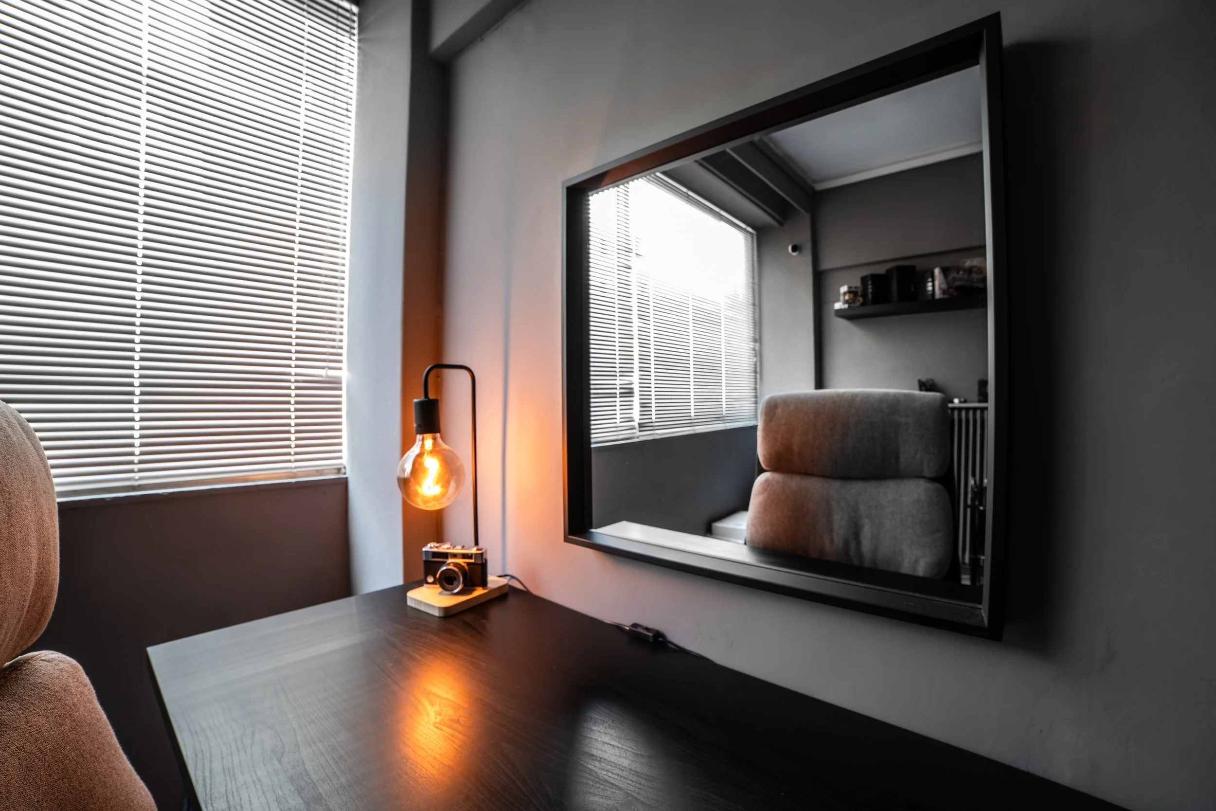 Interior of a room with a wooden desk, a vintage camera on a stand, a modern lamp, and a large mirror reflecting a window with blinds. The walls are painted dark gray, and sunlight filters through the blinds.