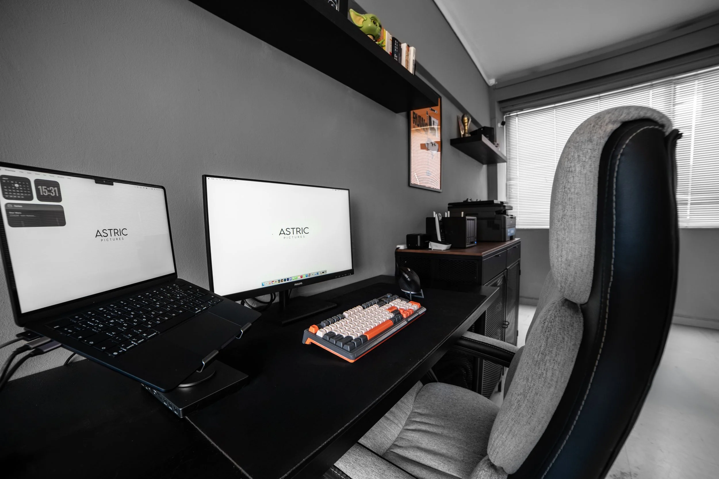 A home office setup with two monitors, a keyboard with orange accents, a laptop, a chair with a high back and gray upholstery, a shelf with books and toys, and a window with blinds.