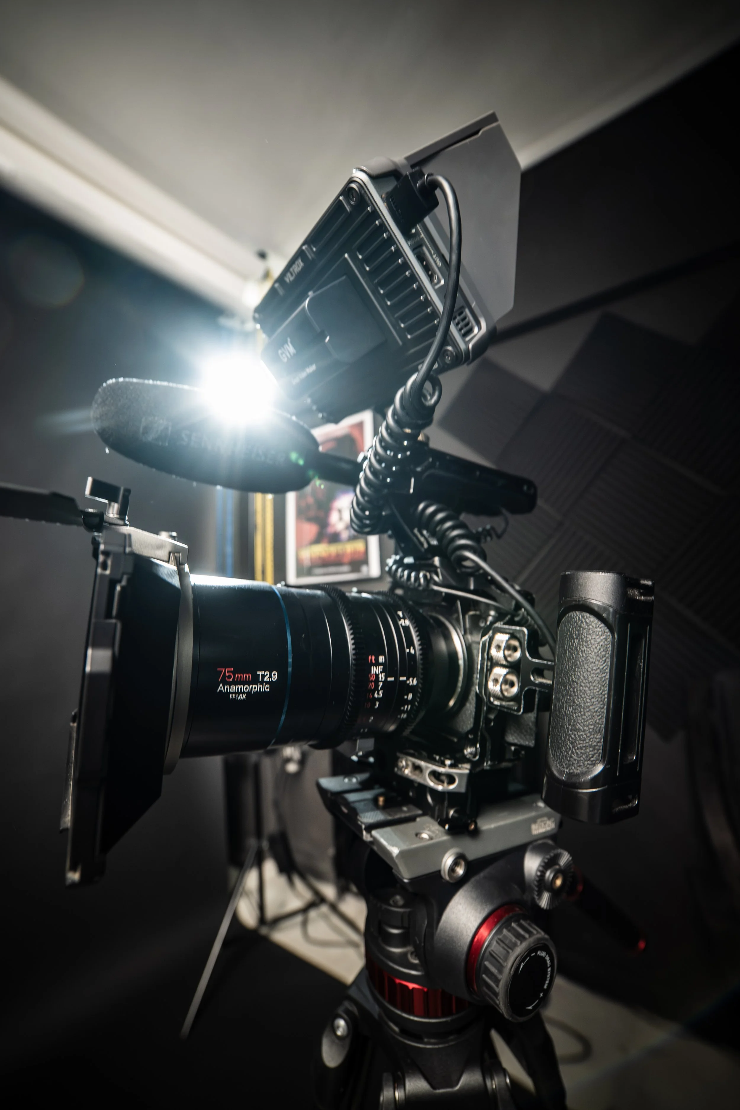 A professional camera mounted on a tripod with a camera mounted on top, positioned indoors with soundproofing panels on the walls. A bright light illuminates the camera setup.