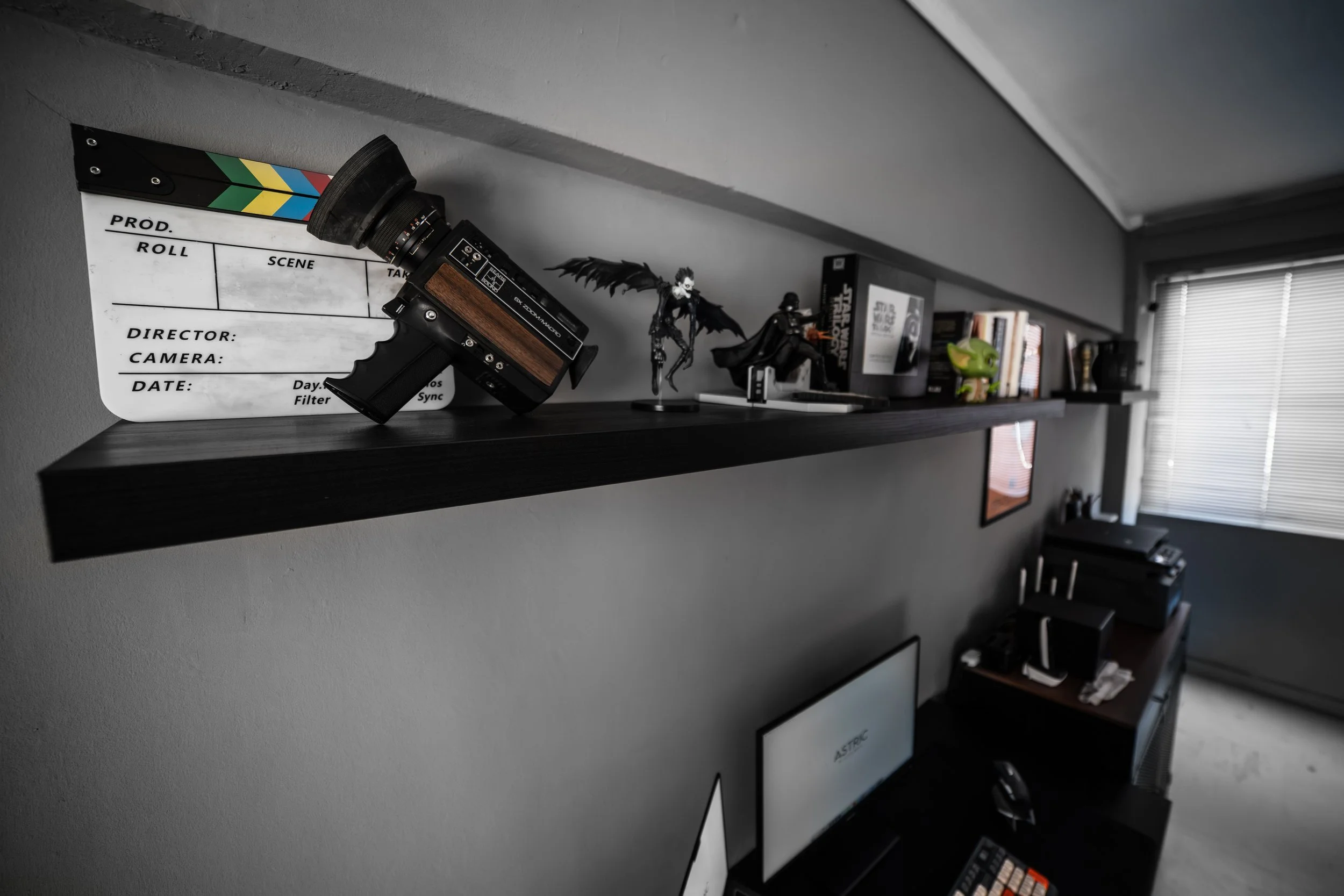 A black shelf against a gray wall displaying collectibles, including a Batman figure, a Star Wars Darth Vader figurine, and a green piggy bank, with books and a window with blinds in the background.