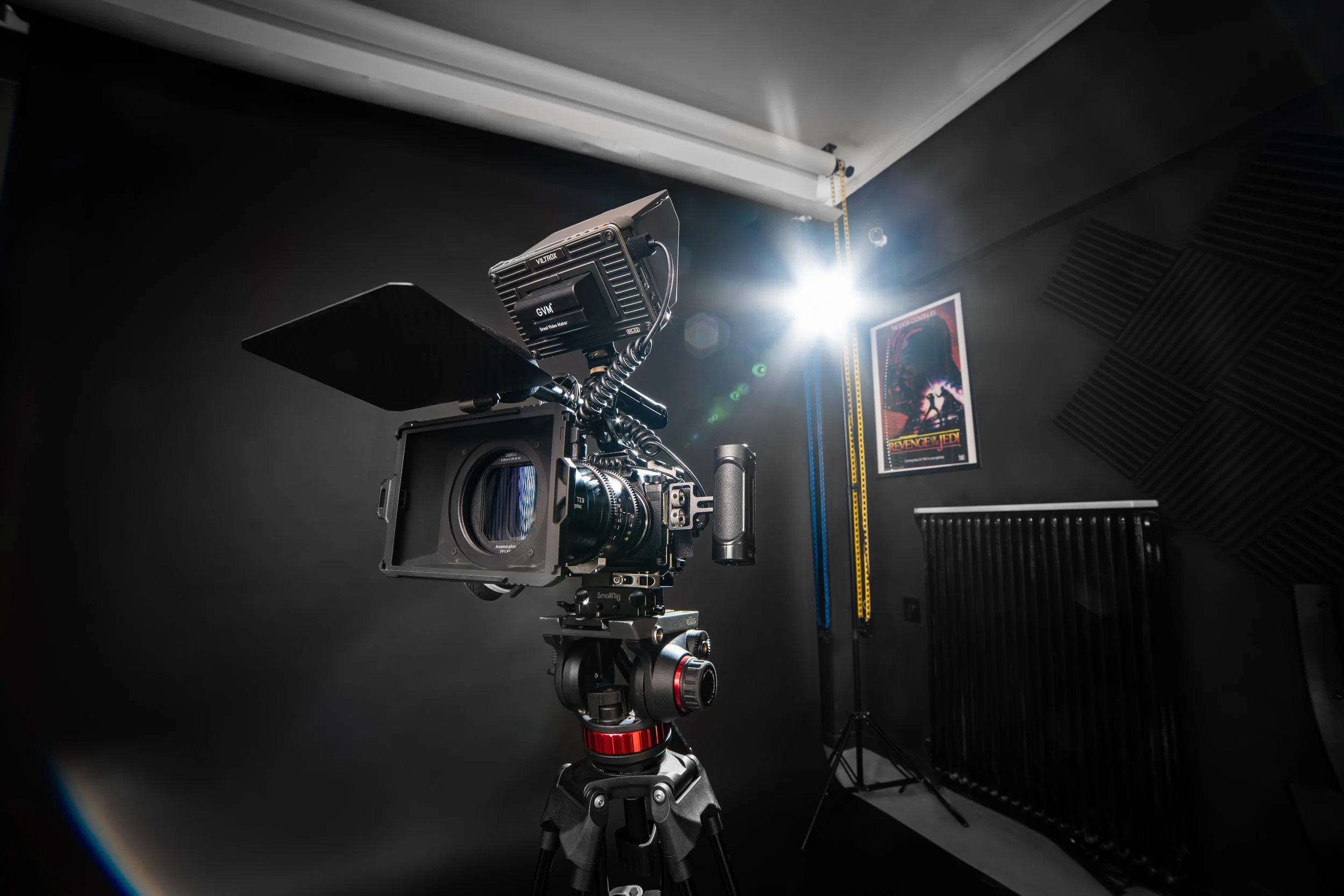 A professional camera mounted on a tripod in a film or photography studio, with a spotlight and a movie poster on the wall.
