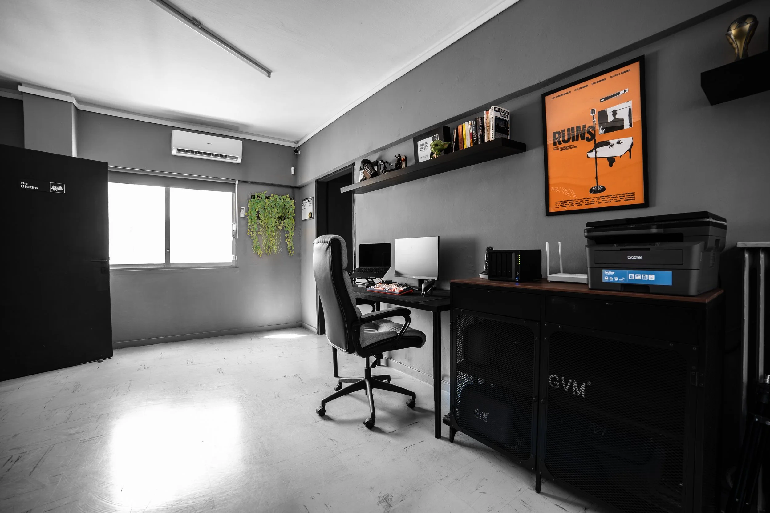 Modern office space with a desk, office chair, dual monitors, printer, bookshelf with books and collectibles, window with blinds, air conditioning unit, and a framed poster on the wall.