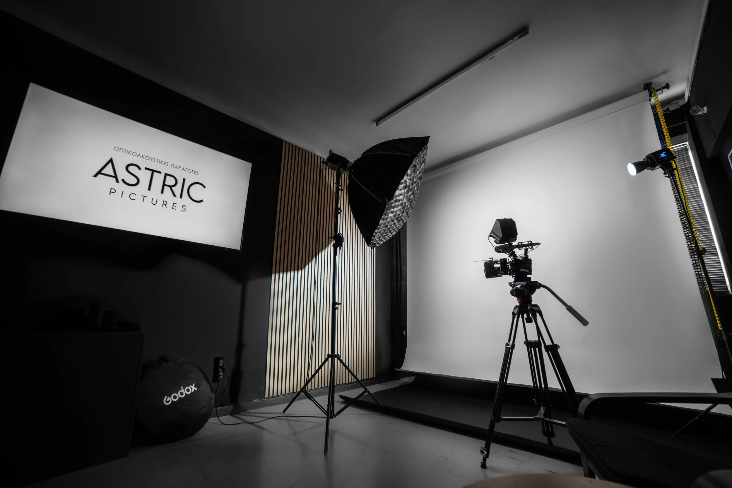 Photography studio with professional camera, softbox lighting, and backdrop, featuring a screen displaying the words 'Optikoakustikes Paragores ASTRIC Pictures.'
