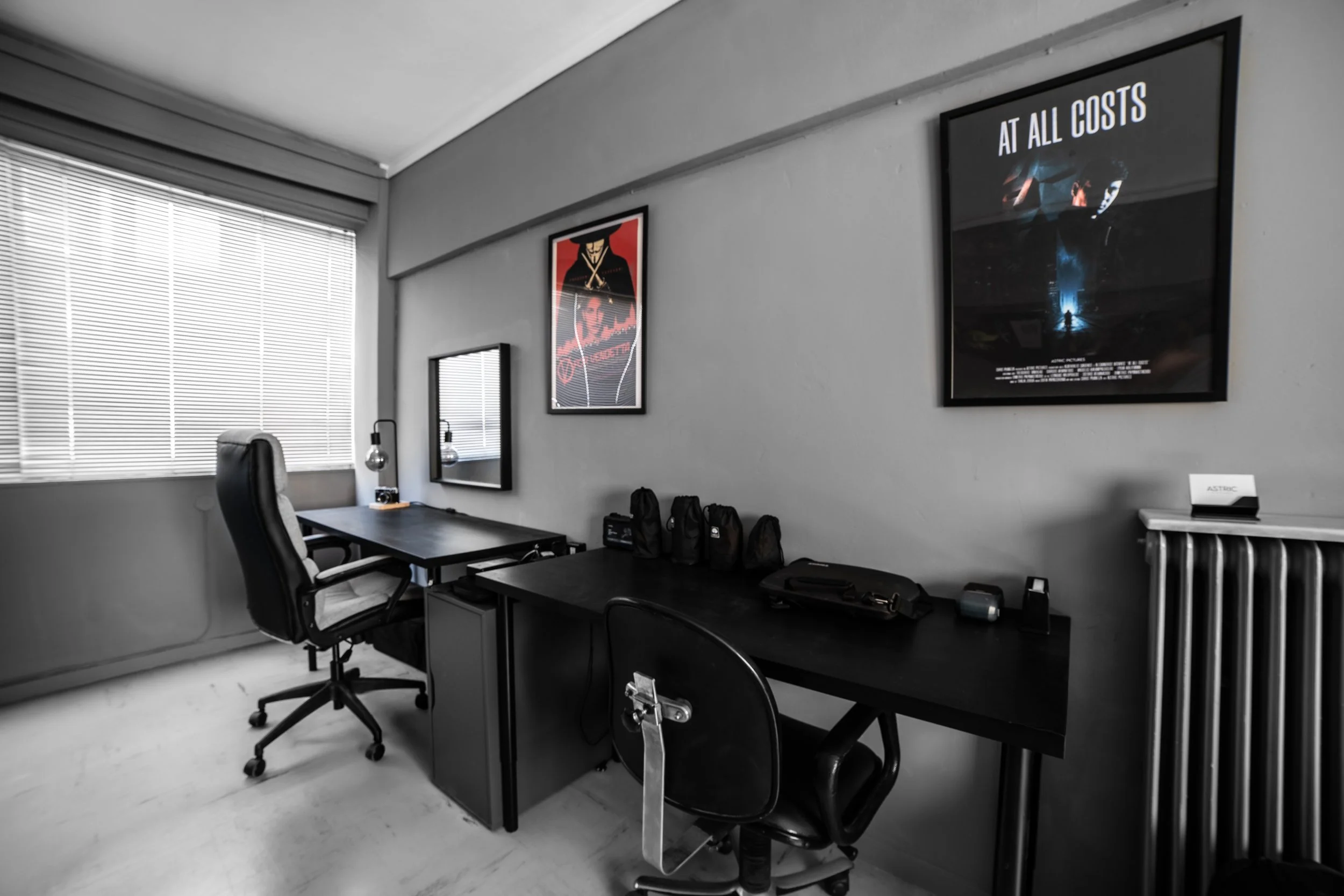 Office room with a desk, black office chair, and various black backpacks or bags on the desk. There are framed posters on the wall, including one with the text "AT ALL COSTS" and an image of a soldier. The room has large windows with blinds and a rad