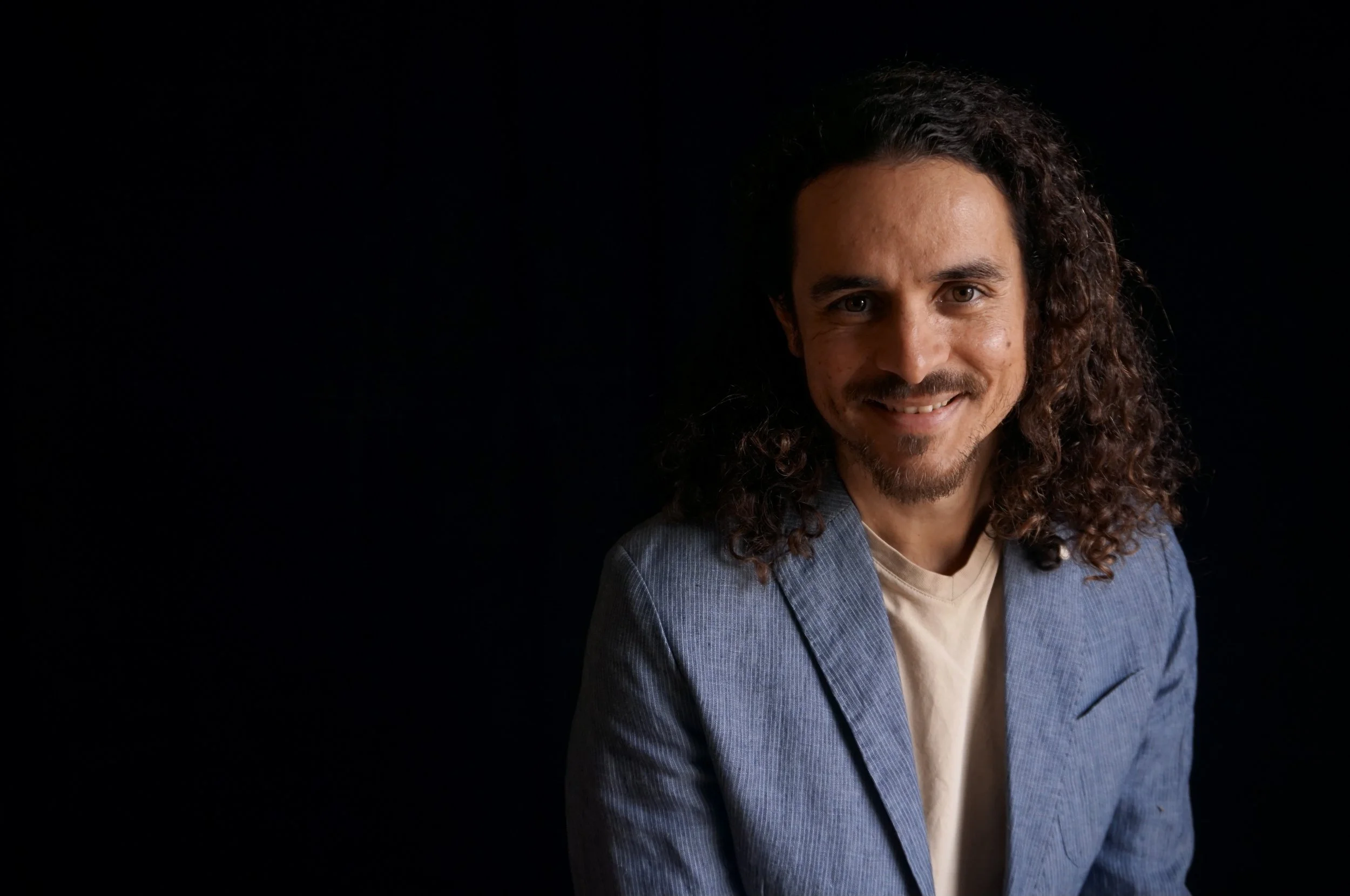 A man with curly hair and a beard smiling, wearing a light-colored shirt and a blue blazer against a dark background.
