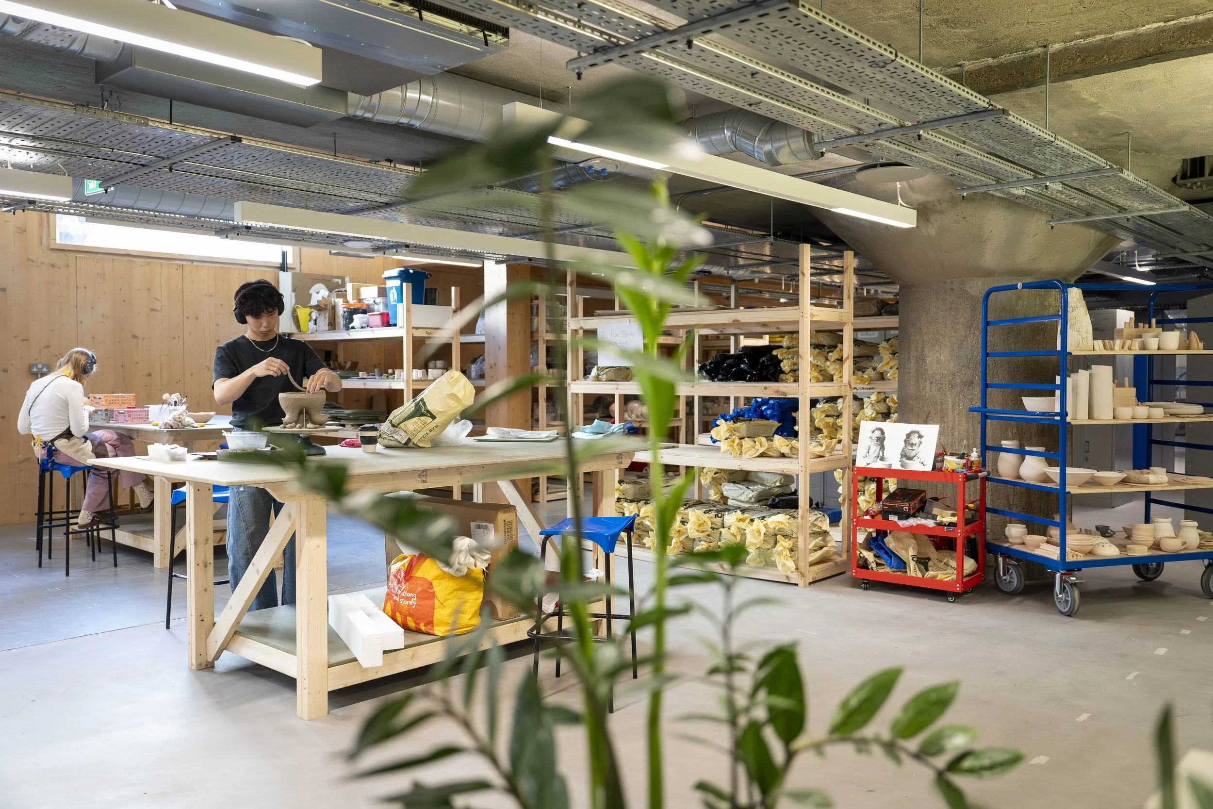 Interior of a bright, spacious pottery studio with shelves full of unglazed pottery, two people working on pottery pieces at a work table, and shelves with pottery supplies.