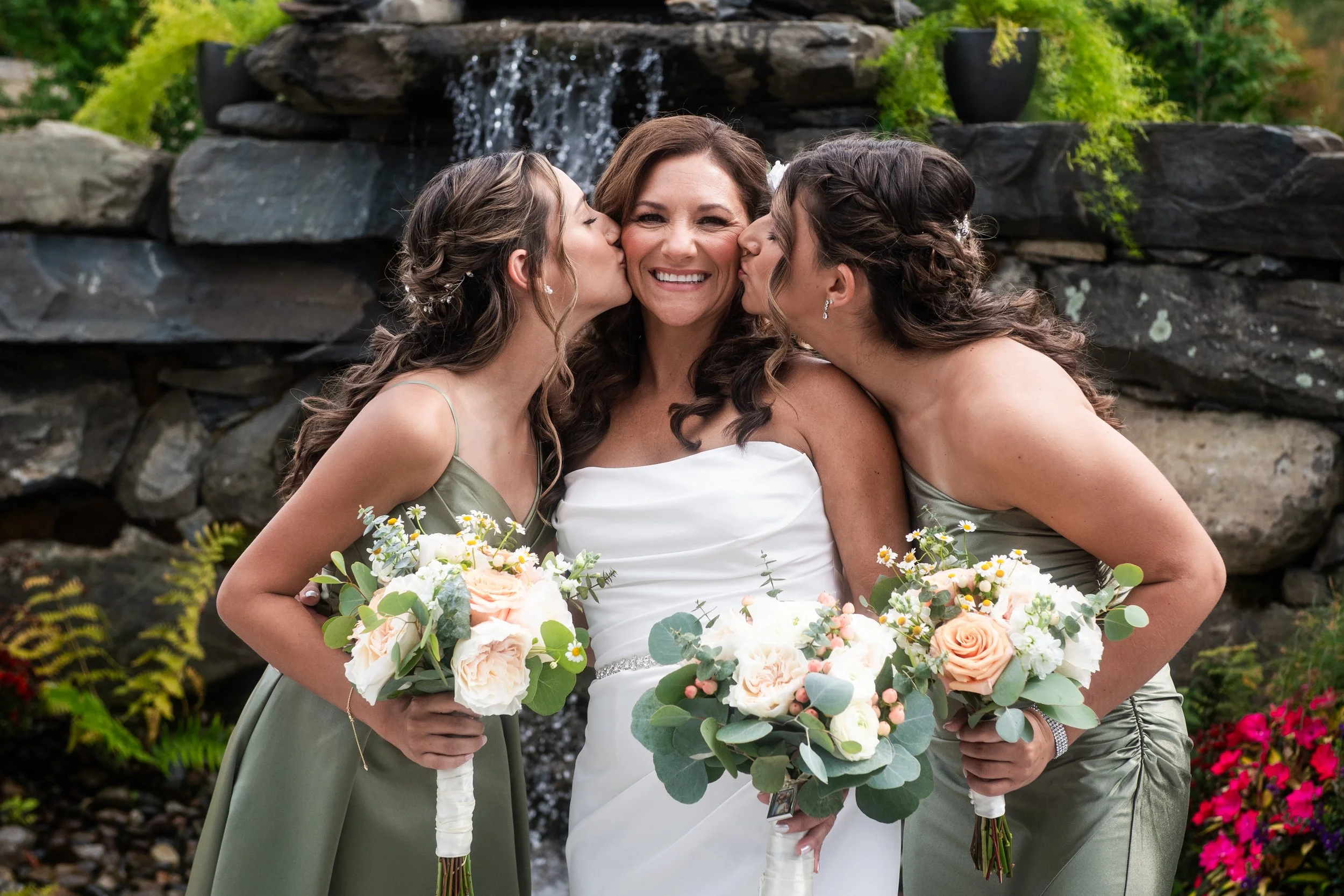Bride smiles as two bridesmaids in sage green dresses kiss her cheeks by the terrace waterfall at Novellas in New Paltz, NY