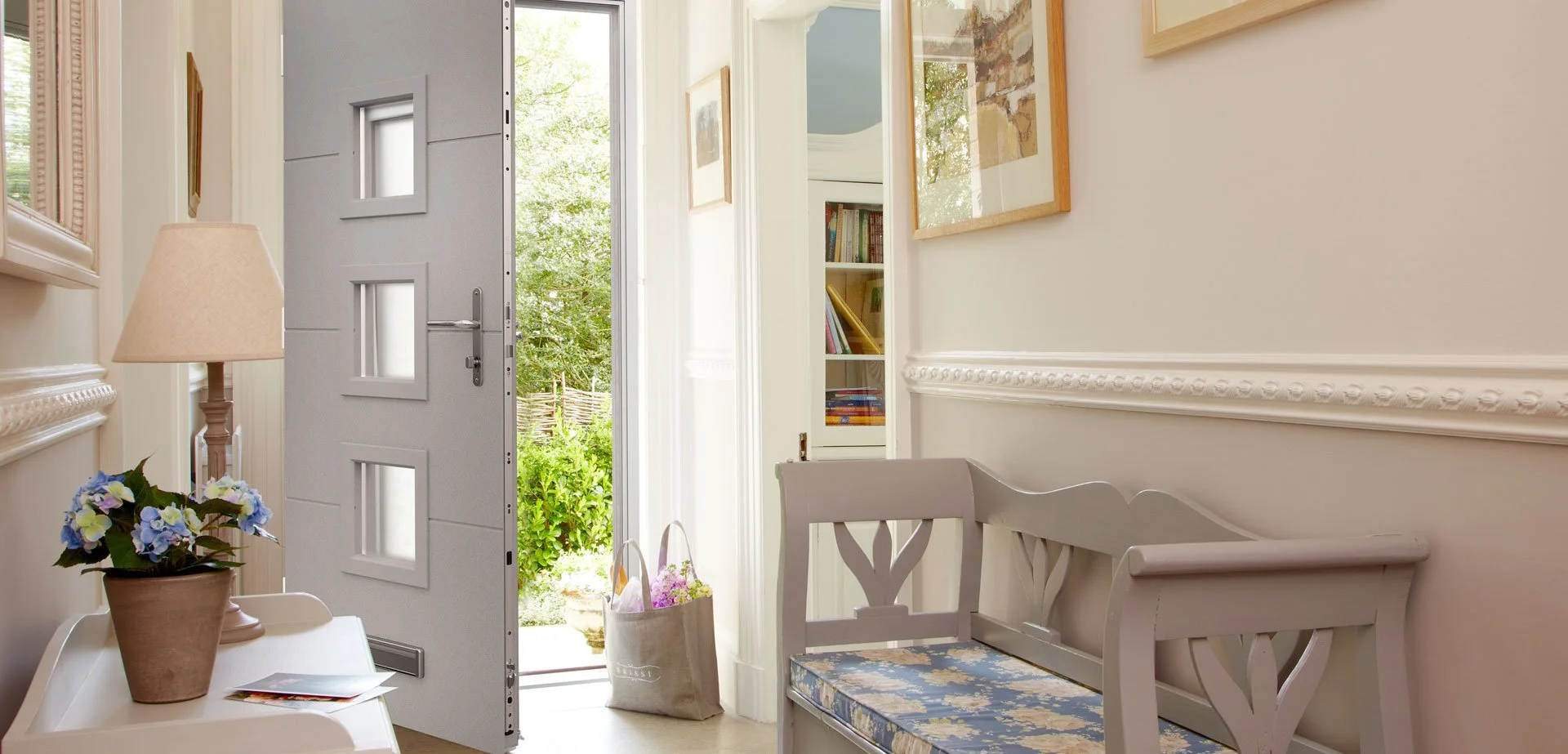 Bright entrance hall with open door leading outside, decorated with a small side table holding a lamp and a potted purple and white flower plant, a bench with floral cushion next to white walls adorned with framed pictures, and a bag filled with flow