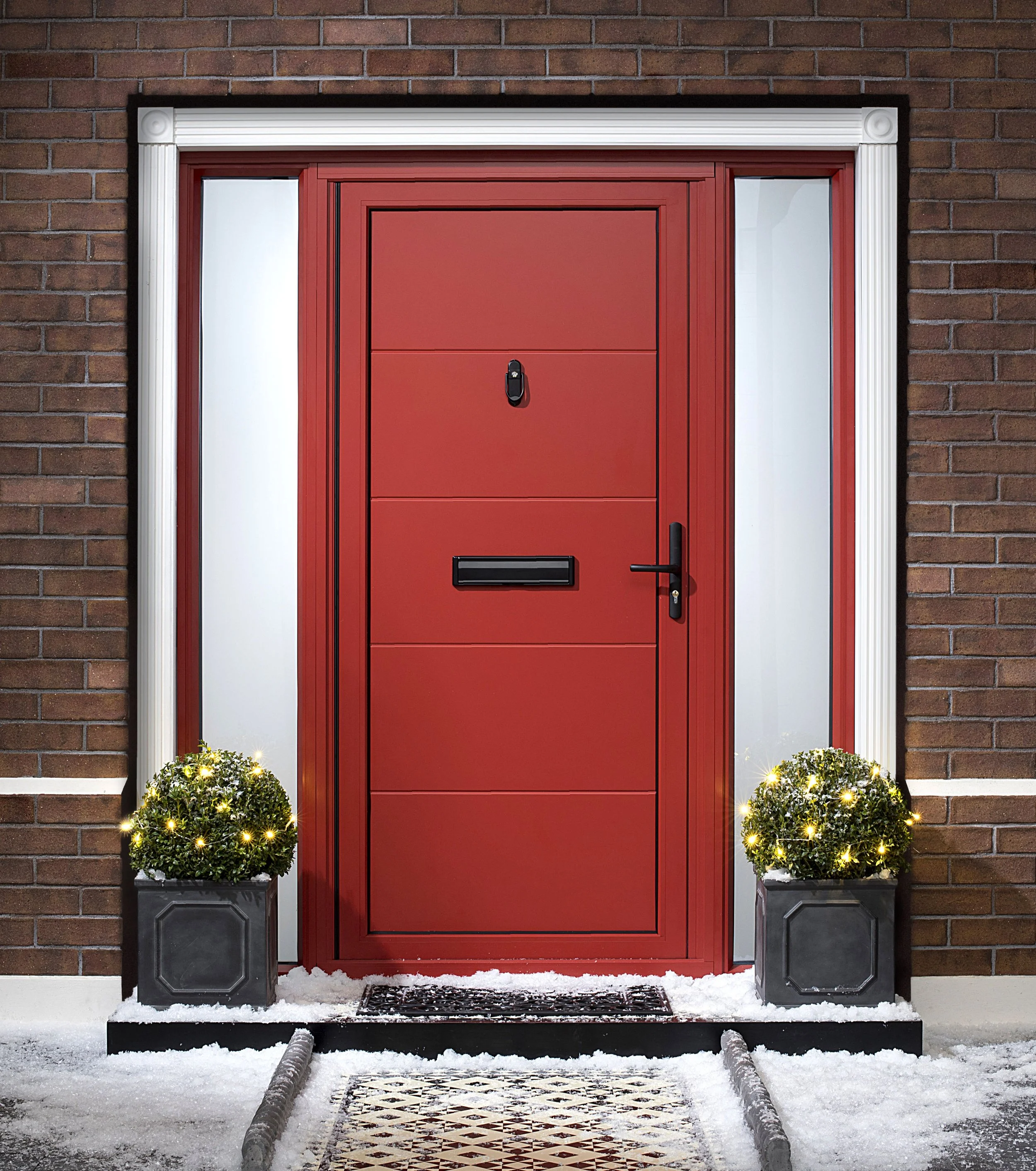 Red front door with black mail slot, handle, lock, and doorbell, framed by white trim, flanked by two potted plants with small yellow lights, on snow-covered ground, against a brick wall.