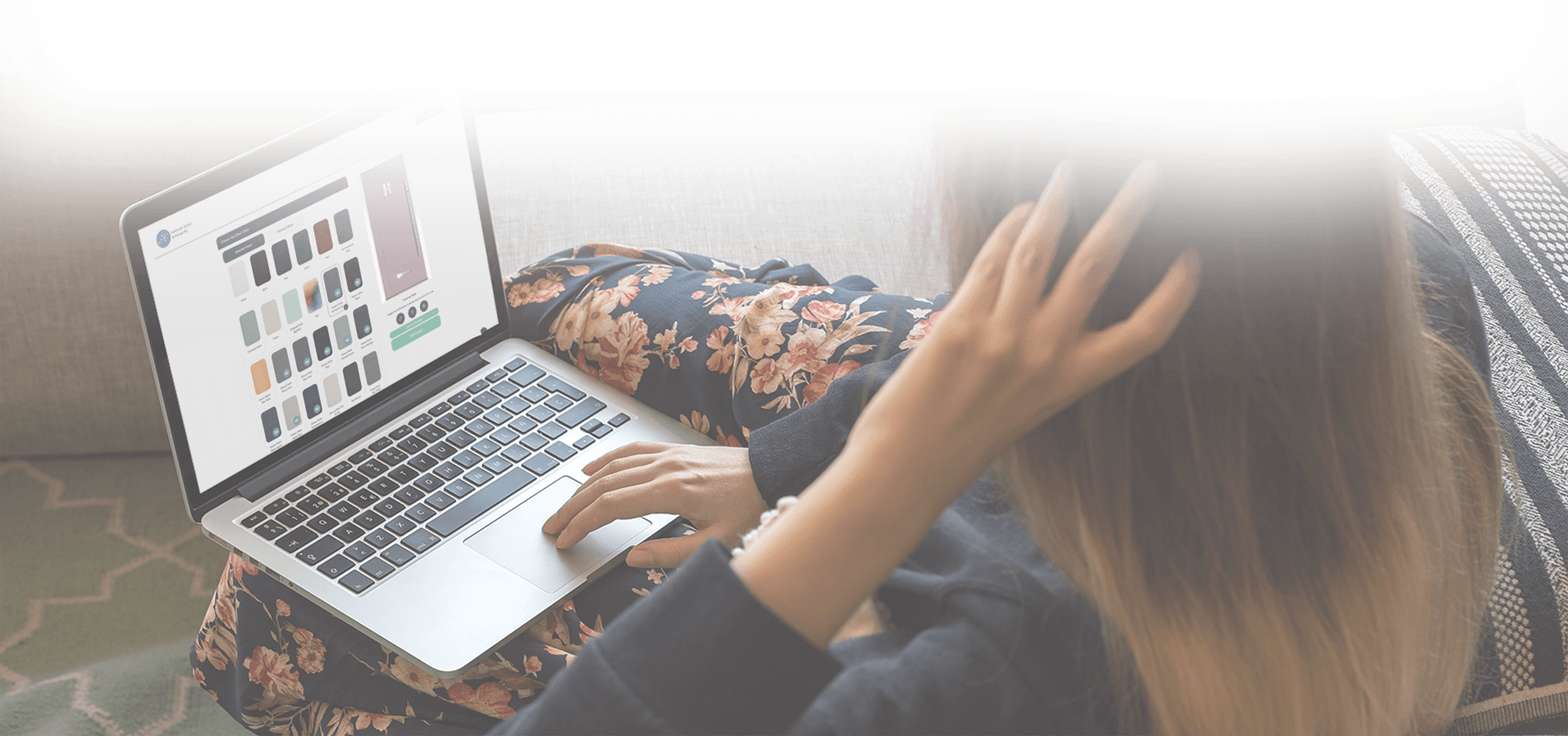 Person sitting on a couch using a laptop, adjusting hair with one hand, with a floral blouse and striped cushion visible.