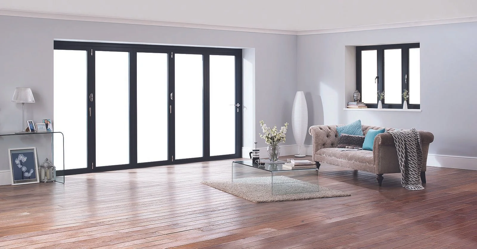 Bright living room with large black framed sliding glass doors, a beige tufted sofa with decorative pillows, a glass coffee table with a plant and books, and a window with decorative items inside on the sill.