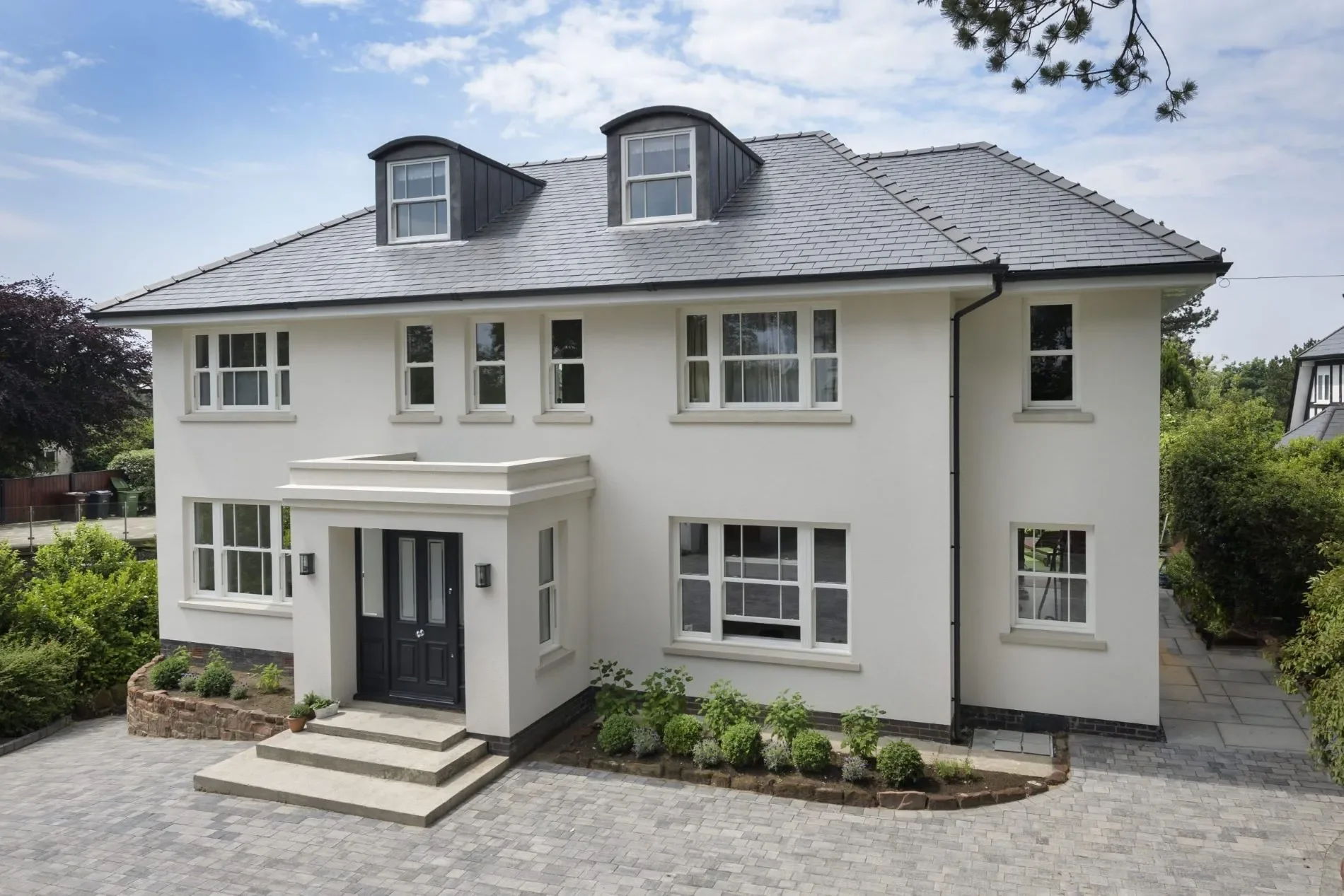 White modern house with multiple windows, black front door, and a gray tiled roof with two dormer windows. The front yard has a paved pathway and small landscaped garden with shrubs and plants.