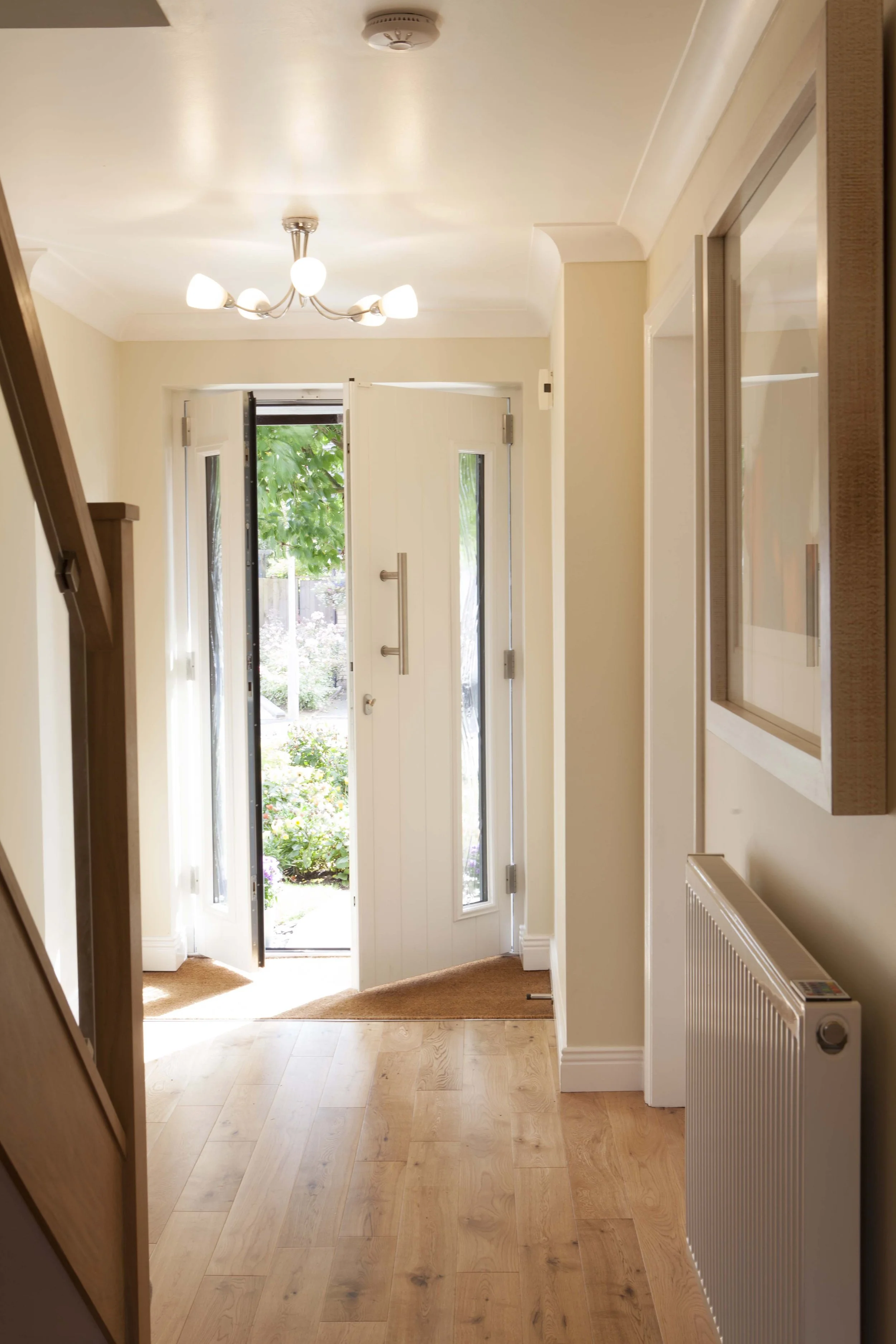 View of a bright hallway leading to a front door with glass panels, with a view outside of green trees and flowers, wooden flooring, a wall-mounted radiator, and a ceiling light fixture.