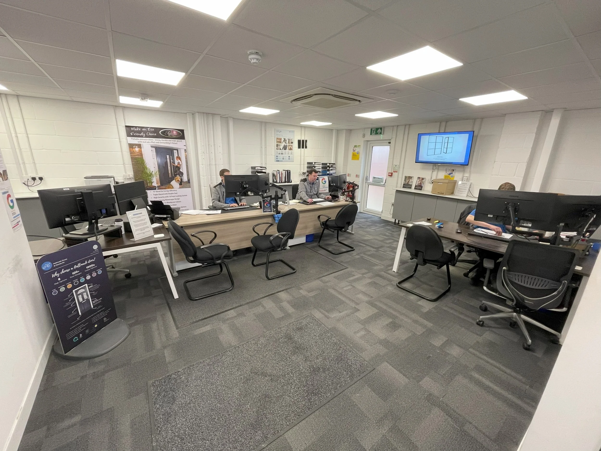 An office with multiple desks and chairs, computer monitors, and a flat screen TV mounted on the wall. There are people working at their desks.