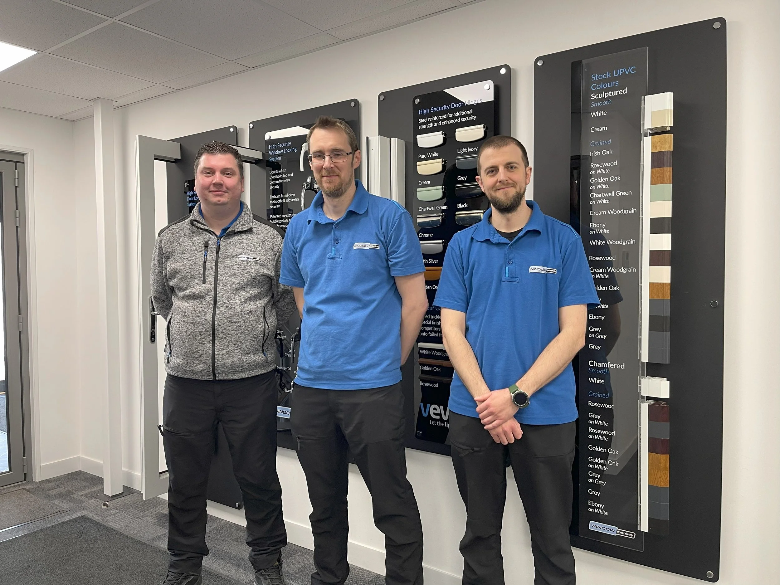 Three men standing in front of display boards showing different colors and styles of UPVC window frames and finishes in a showroom or office.