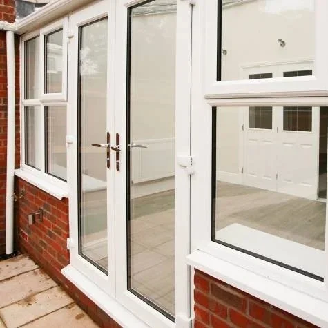 Photo of a large glass door with white framing, leading into an indoor space, with a window to the right, a brick wall base, and a white window visible through the glass.