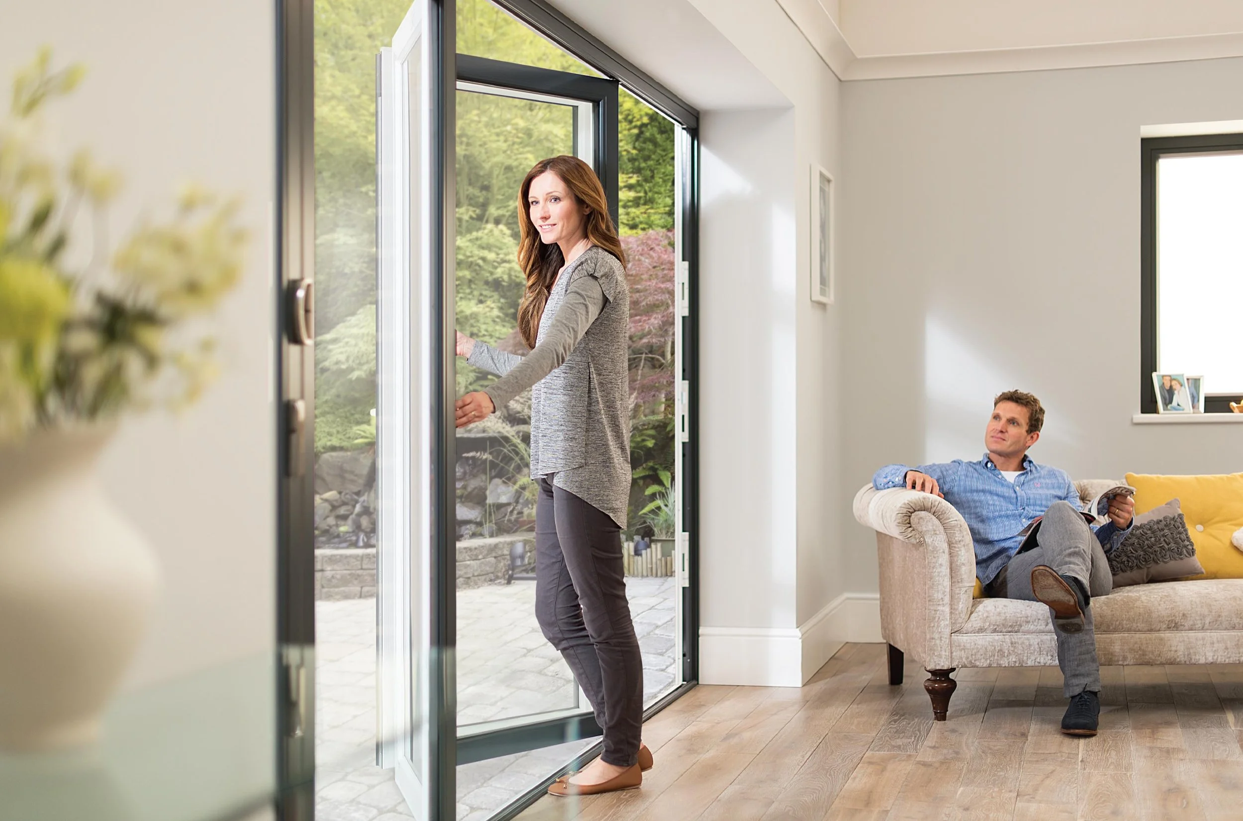 A woman opens a sliding glass door to a patio outside, while a man sits on a beige couch inside the living room, looking at her.