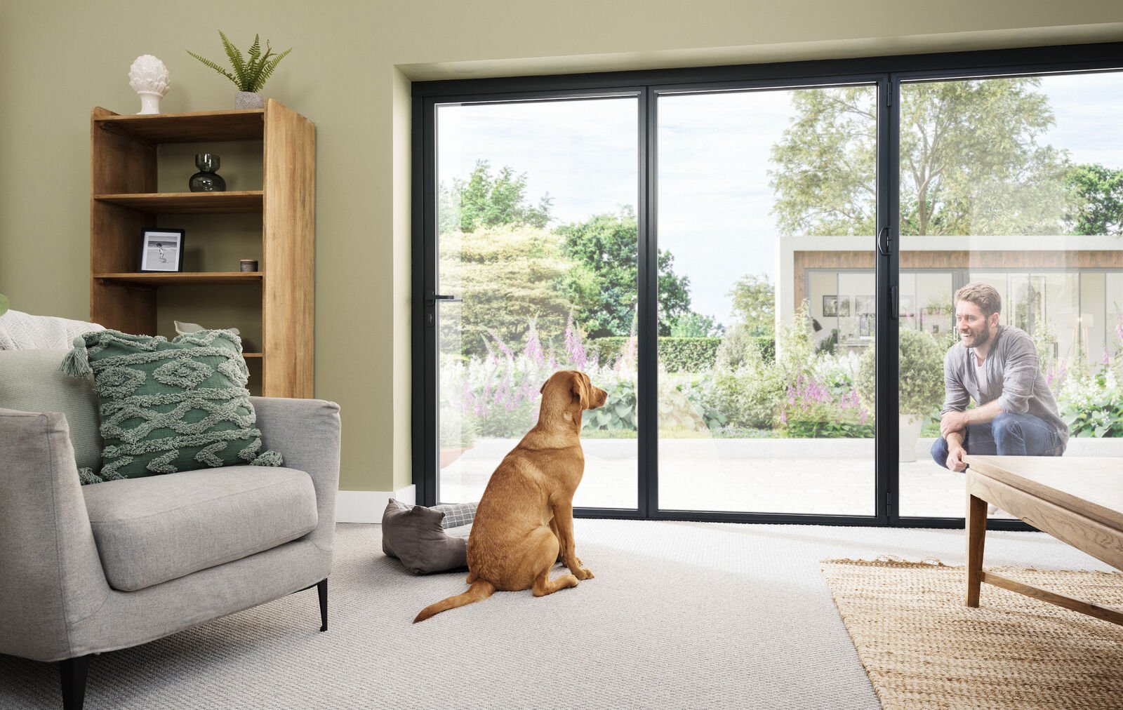 A living room with a gray sofa, decorative pillows, a wooden bookshelf, and a sliding glass door leading outside. Inside, a brown dog sits on the carpet, looking at a man outside through the glass door, who is squatting and smiling.