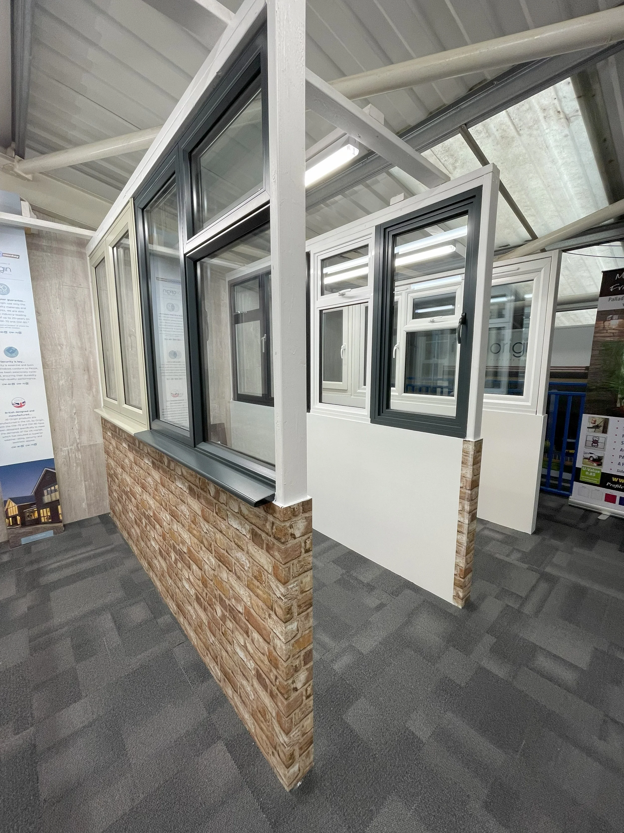 Display of various modern window frames in a showroom, featuring different colors and styles, with brick and white wall backgrounds.
