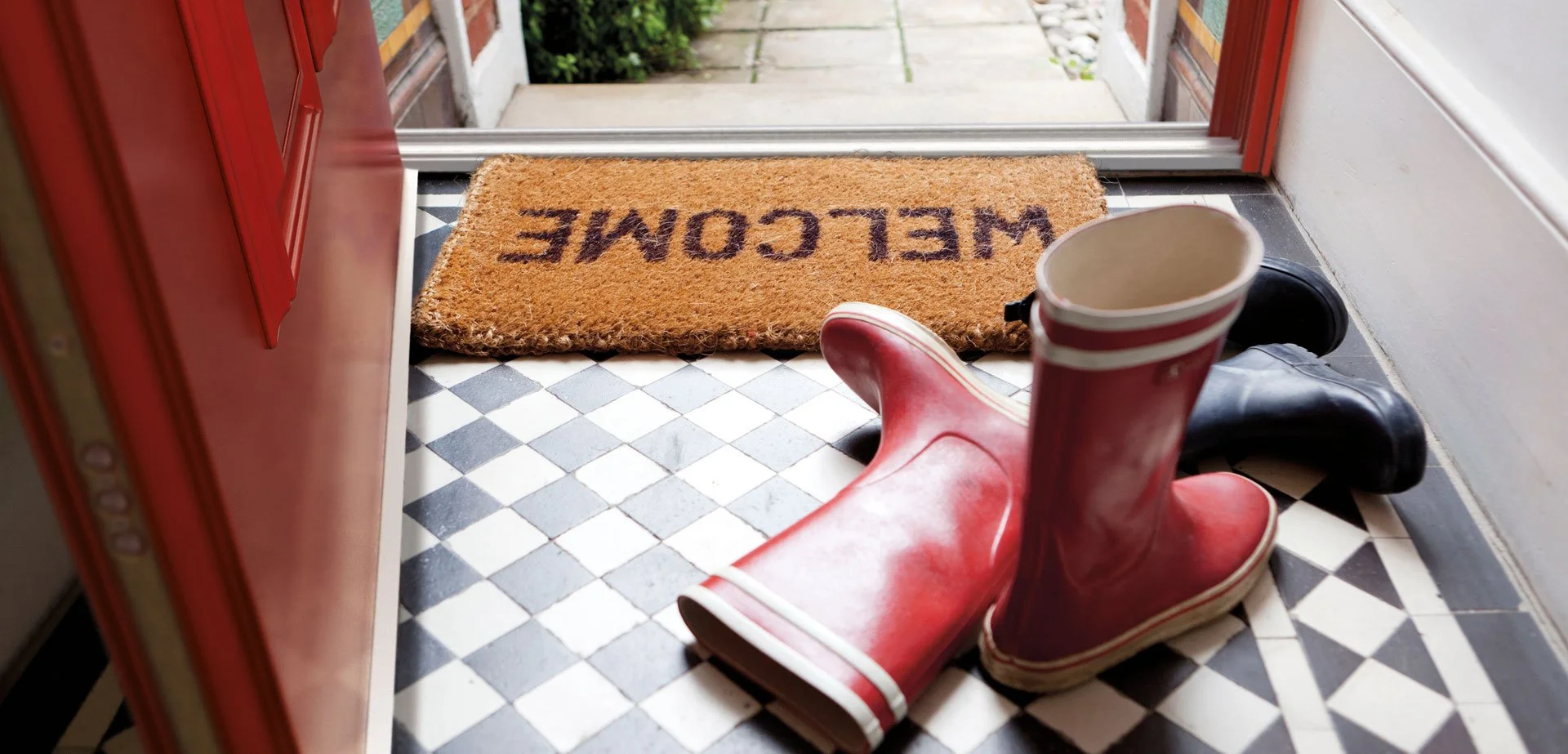 A welcoming front door entrance with a doormat that says "WELCOME". On the black and white checkered floor, there are two pairs of rubber rain boots, one red and one black, with the red pair positioned on top of the black pair. There is also a white 