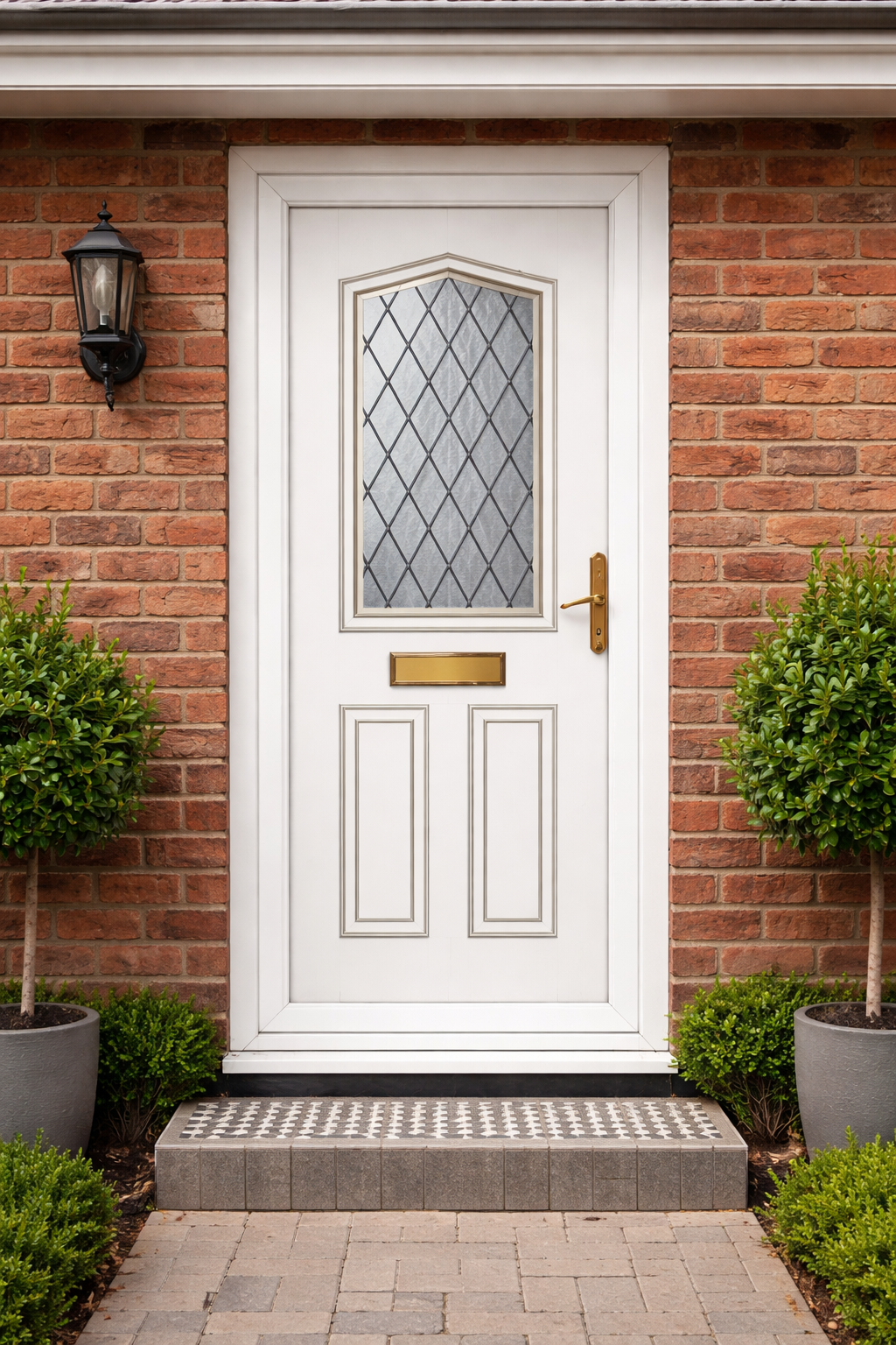 White front door with diamond-patterned glass window, brass handle, mail slot, surrounded by brick wall, flanked by green shrubs in pots, with outdoor lantern on the left