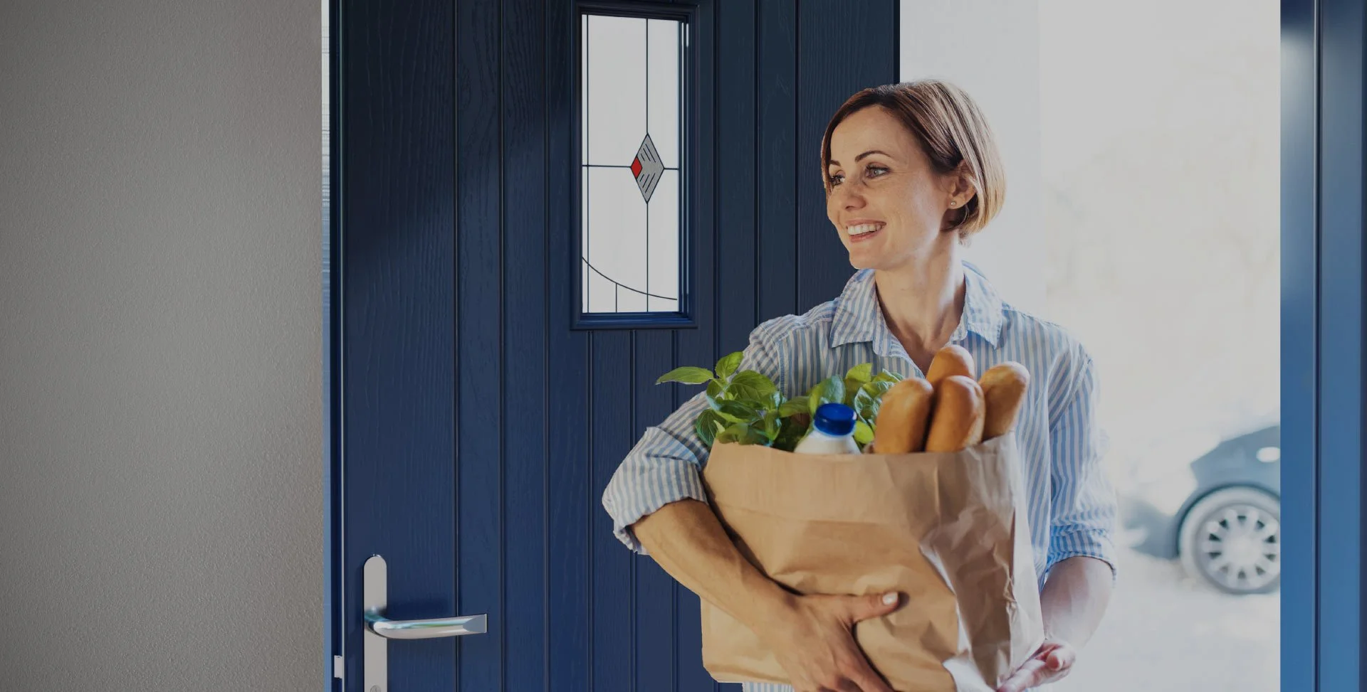 A woman with short hair smiling as she walks through a blue door holding a paper grocery bag filled with bread, a plastic bottle, and green leafy vegetables.