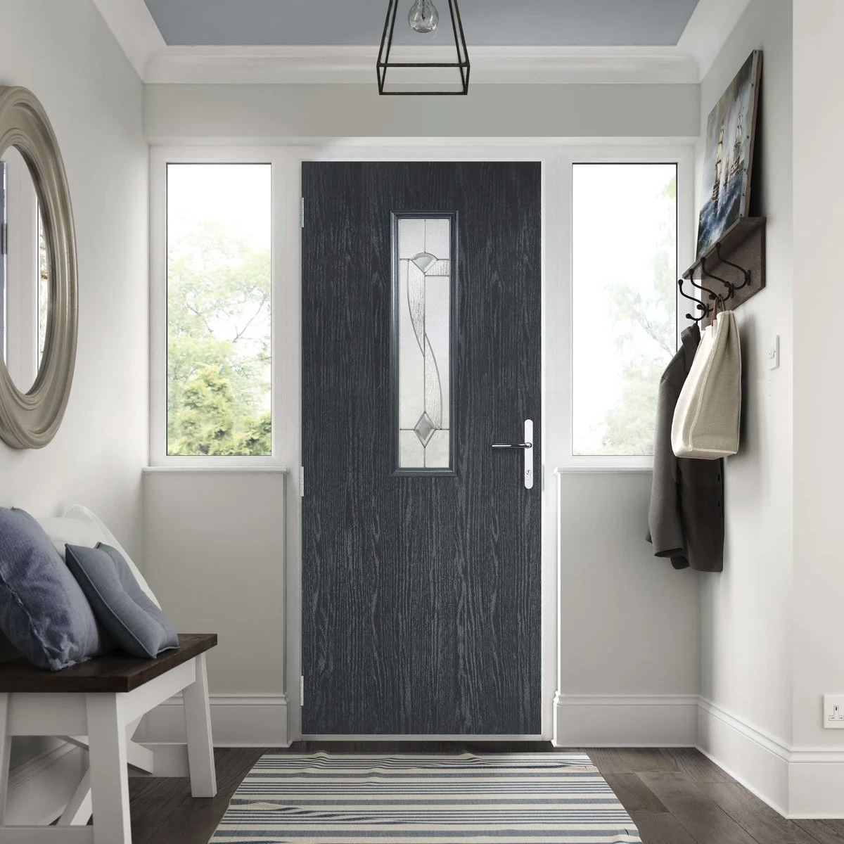 Interior view of a modern entryway with a black front door featuring a decorative glass panel, flanked by two windows, a bench with pillows on the left, and a wall-mounted coat rack holding jackets and a tote bag on the right, with a striped rug on t
