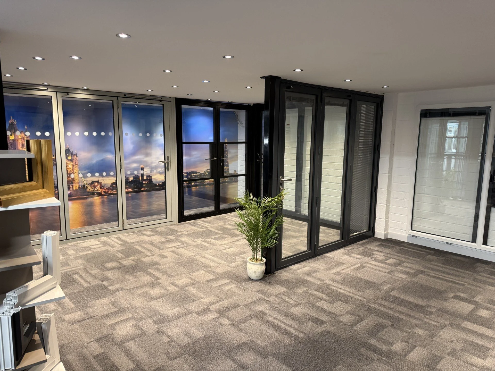 Showroom space with bifolding doors showing a cityscape during sunset, with a carpeted floor, a potted plant, and a shelving unit. The ceiling has multiple small recessed lights.