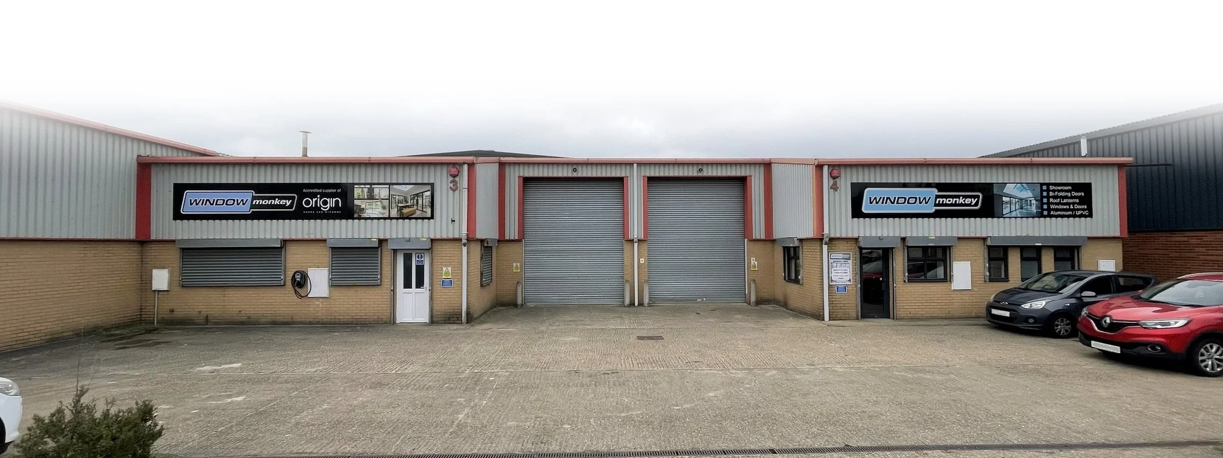 Exterior of a commercial building with parking lot, two signs for Window Monkey, roller shutters, and several parked cars.