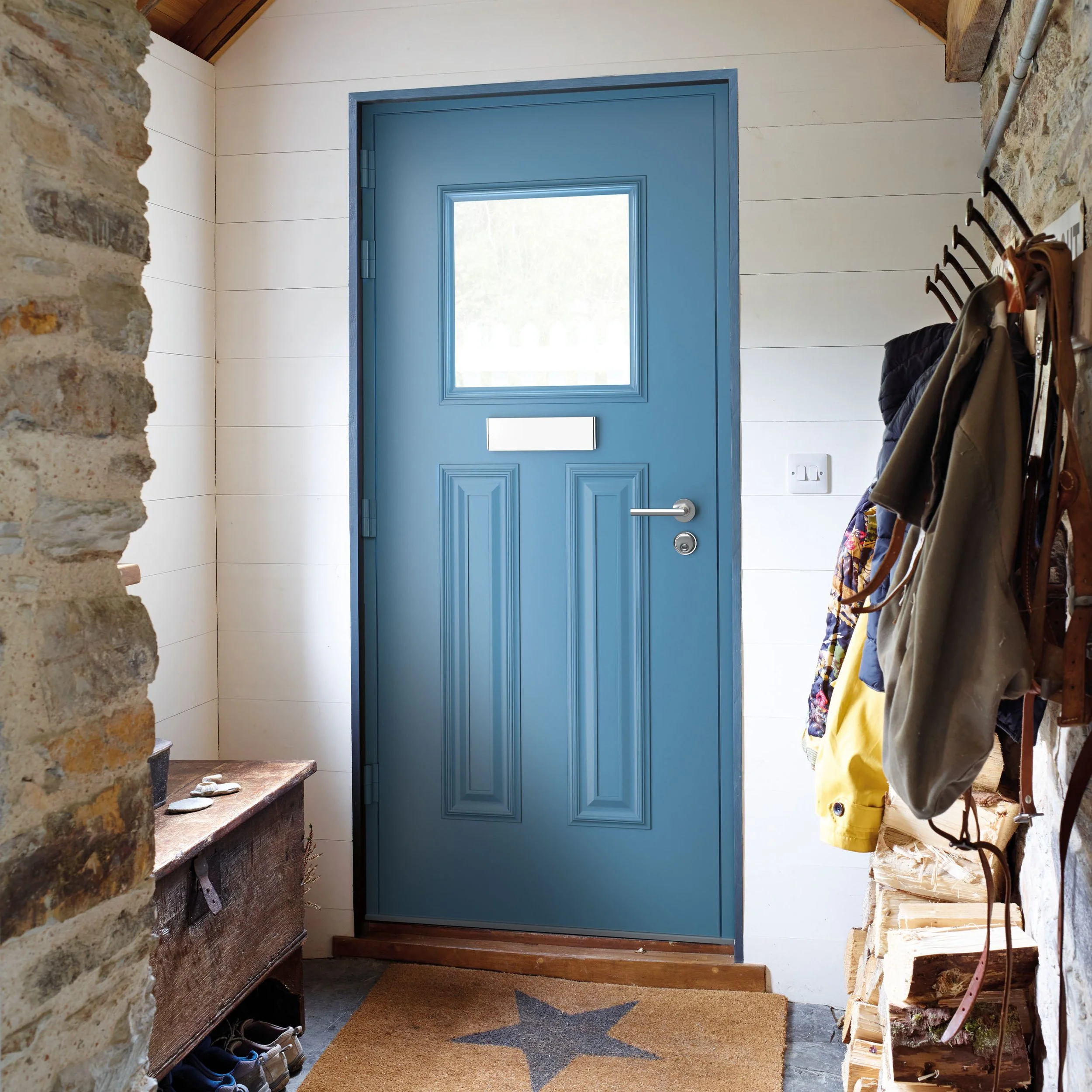 Blue front door with a letterplate and window, set in a small entryway with a star-shaped rug on the floor, wooden logs on the right, and a vintage wooden chest on the left, with coat hooks holding jackets and bags.