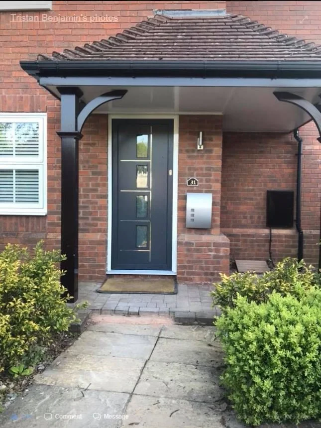 Front porch of a brick house with a black door, a mailbox, and a small covered area supported by black columns, with shrubbery on either side and a concrete walkway.