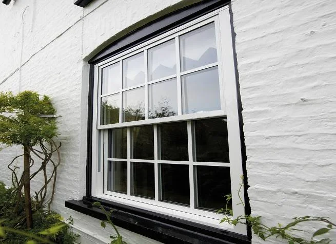 A white house with a double-hung window featuring a black trim and a black window sill, surrounded by green plants and shrubs.
